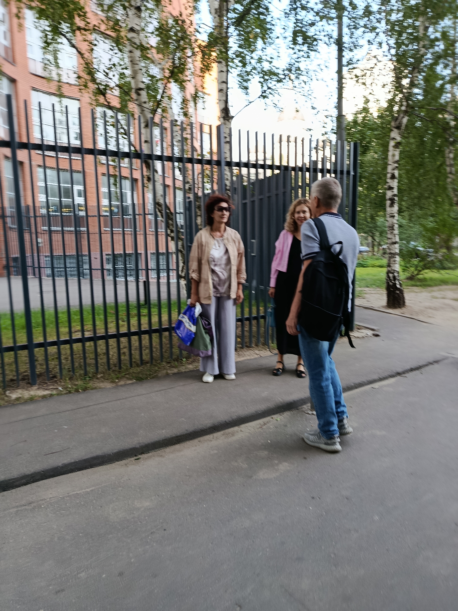 In Moscow, Russia, on a paved pathway adjacent to a black metal fence, three individuals are present. A woman in the foreground, facing away from the camera and towards a red brick building with numerous windows, is dressed in a light-colored jacket and gray pants. She carries a blue and white shopping bag and a green bag. To her left, a woman in a pink jacket over a black dress is visible, with a black bag at her feet. To the right, a man wearing a grey polo shirt, blue jeans, and a black backpack is facing the two women. The scene is set outdoors with trees and greenery surrounding the area.