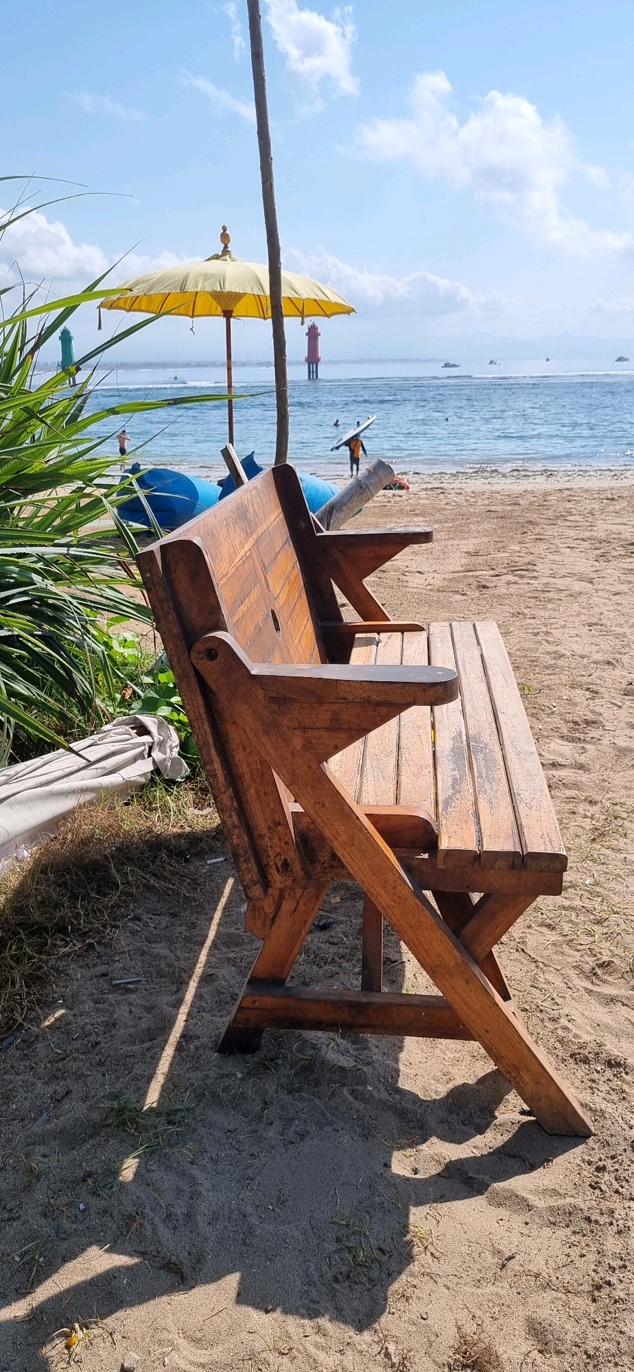 A wooden bench sits on a sandy beach in Denpasar, Indonesia, facing the ocean. The bench is empty and positioned slightly to the right of the frame, angled towards the water. Behind the bench, lush green foliage is visible on the left side of the image. Further back, a yellow umbrella stands tall, casting a shadow on the sand. The ocean stretches out in the background, with several people in the water and a person carrying a surfboard on the shore. Two lighthouses, one red and one green, stand in the distance on the water. The sky above is a clear blue with scattered white clouds. The scene suggests a peaceful day at the beach, with the bench offering a place to relax and enjoy the view.
