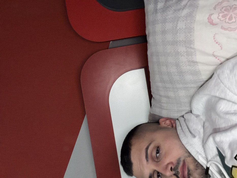 The scene depicts an adult male in an indoor setting, identified as Opanets, Bulgaria. The individual is positioned lying down, with his head resting on a pillow. He possesses short dark hair with a fade haircut and a light beard or stubble on his face. He is wearing a light grey hooded sweatshirt. The pillow features a grey and white checkered pattern on its upper half, transitioning to a white lower half with a subtle pink floral motif. Behind the subject's head, a modern headboard is visible, characterized by a predominantly red color scheme with distinct curved white sections and additional dark red elements. A solid dark red surface occupies the upper portion of the frame. The male's gaze is directed towards the camera.