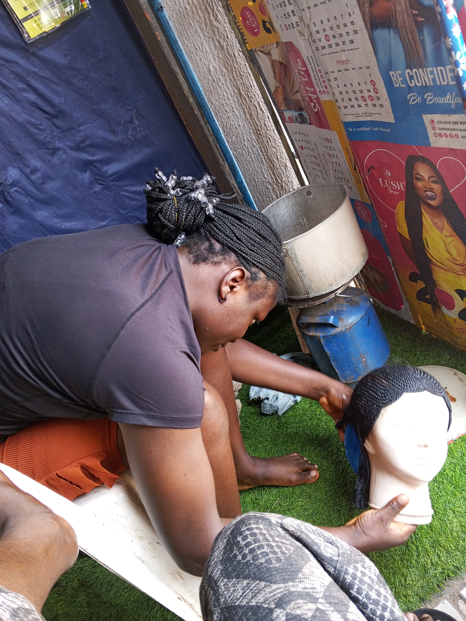 A dark-skinned person with braids is seated outside on a white chair. The person is wearing a black t-shirt and an orange skirt. They are holding a wig on a mannequin head. Their bare feet are visible on the green artificial turf. The location is a temporary outdoor workspace, with a blue tarp visible above and a stack of items, including a metal pot and a blue container, next to them. Behind these items are calendar posters with images of women and text promoting Lush Hair, including contact information such as phone numbers and social media handles, indicative of a business location, possibly in Gwong, Nigeria. Another person wearing gray patterned pants is partially visible on the bottom right.