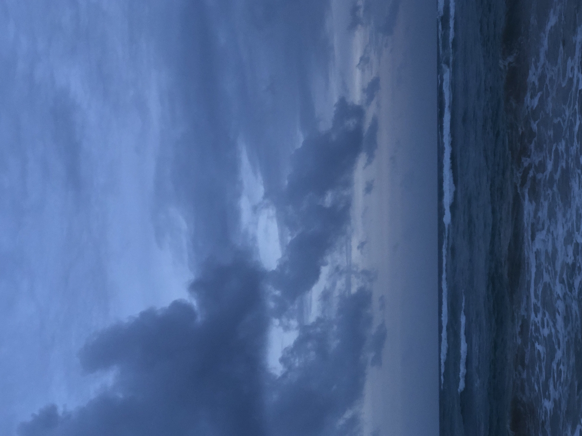 The image depicts a gloomy seascape at dusk in Kathaluwa, Sri Lanka. The sky is overcast with heavy, dark blue clouds, suggesting an impending storm or the remnants of one. The sea is a deep, murky blue, with white-capped waves rolling towards the shore. The foreground is filled with foamy surf, indicating rougher waters closer to the beach. There are no people or objects visible in the scene, emphasizing the natural, untamed beauty of the coastline under the dramatic sky.