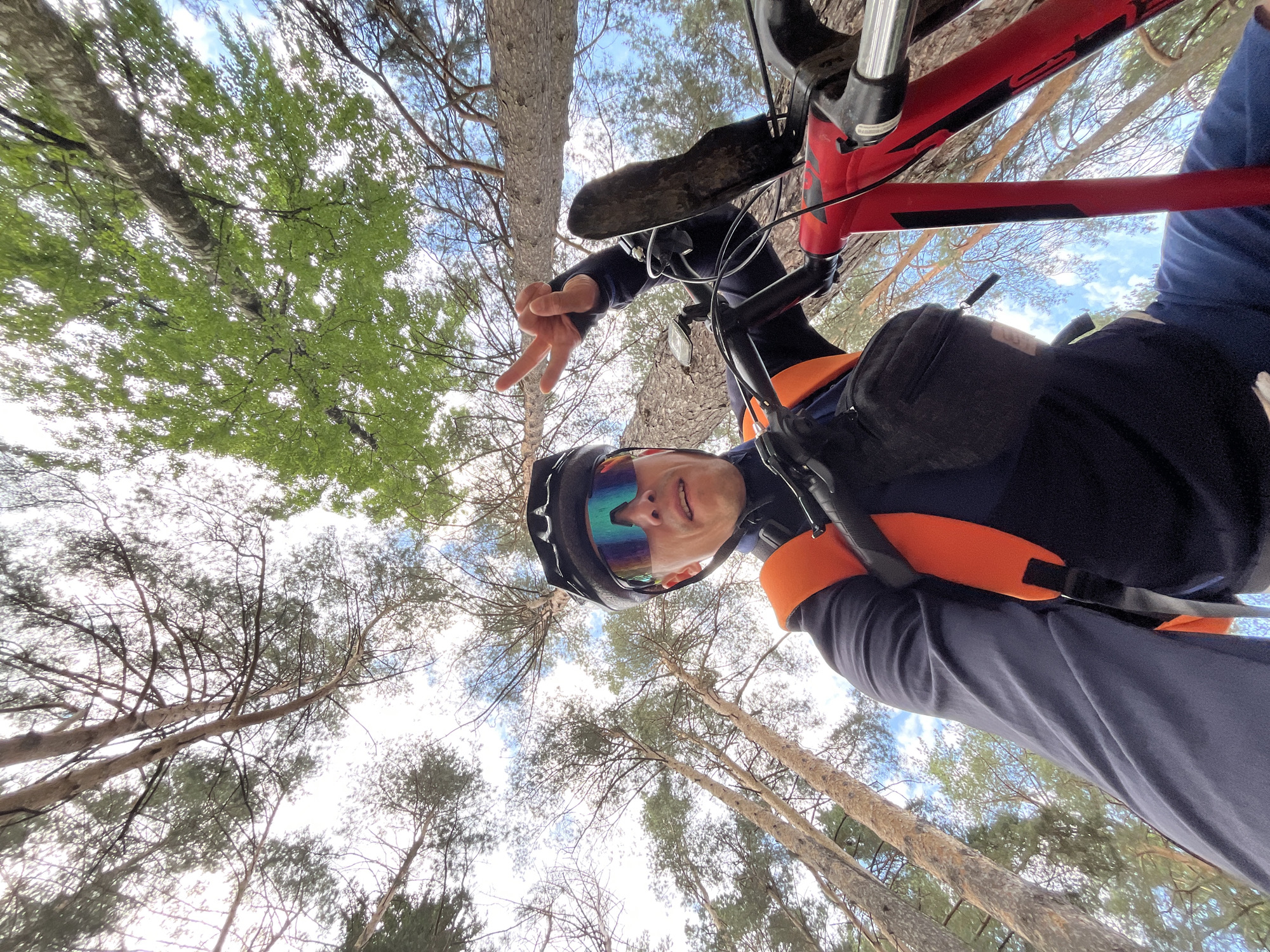 In Chaikovsky, Russia, a person is captured in a low-angle, wide-shot selfie while riding a red mountain bike on a forest trail. The rider, wearing a black helmet, blue long-sleeved jersey, and an orange vest, looks towards the camera, with their left hand raised in a peace sign. The bicycle's handlebars, front suspension, and part of the frame are visible. The background is dominated by tall trees with visible branches and foliage, with patches of blue sky and white clouds peeking through. The forest floor is not visible in this shot.