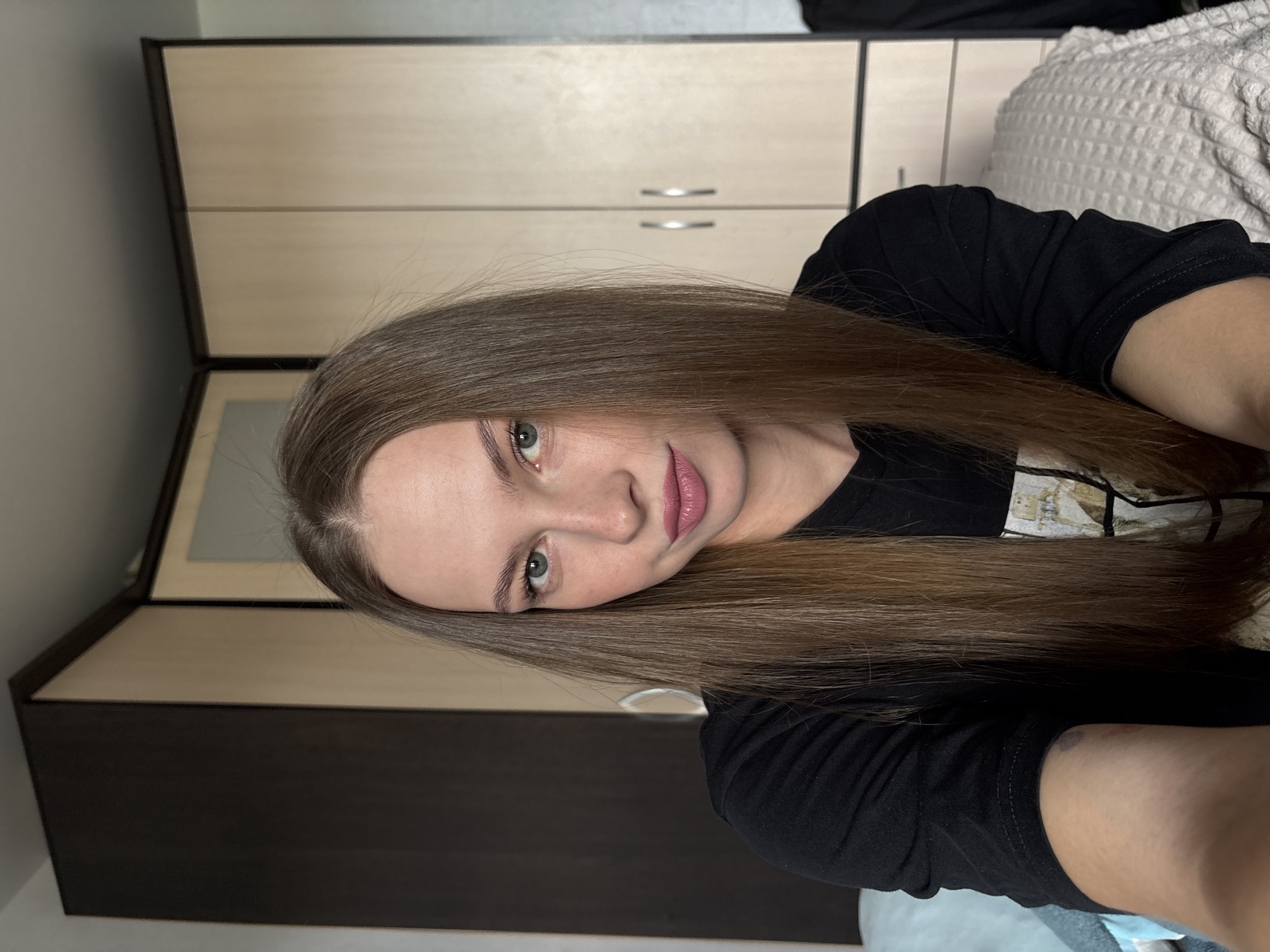 A young Caucasian female with long, straight brown hair, fair skin, and green eyes is captured in a close-up, head-and-shoulders selfie. She is wearing a black t-shirt. Her gaze is directed slightly off-camera. The background suggests an indoor setting with a wardrobe that appears to be a light wood color with dark brown accents. The lighting is soft and natural, indicating it could be daytime. The image does not contain information that would allow for the identification of a specific city like Perm, Russia, or any other location.