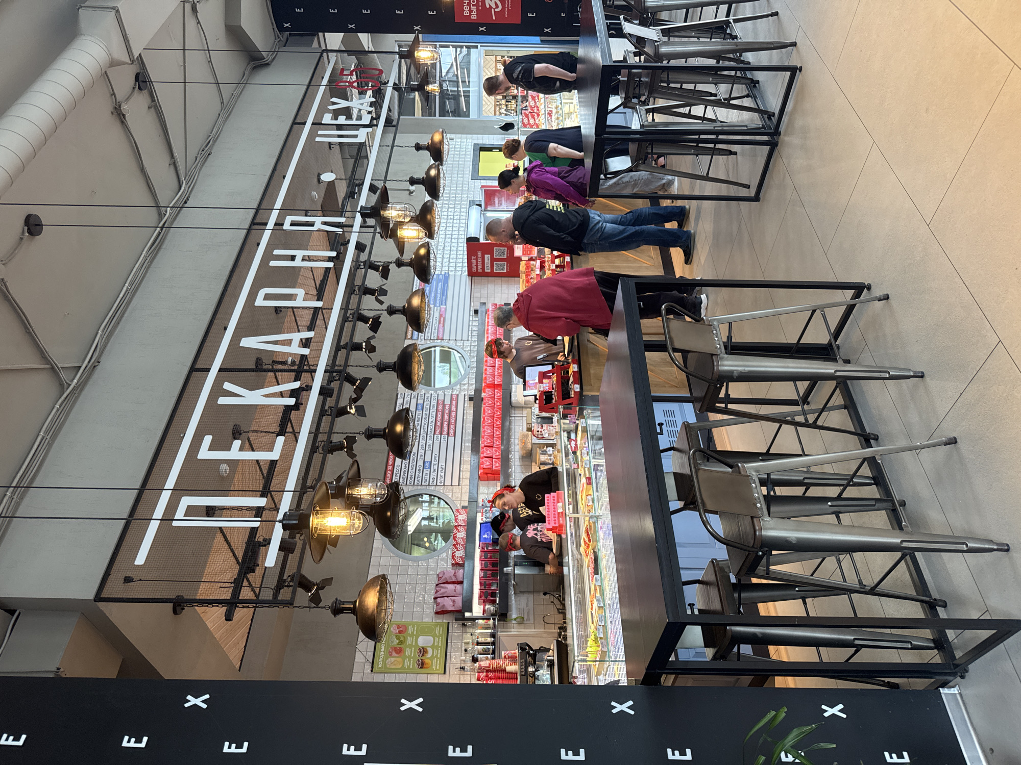 This is a vertical, eye-level shot taken inside a modern bakery or cafe. The scene is brightly lit with overhead industrial-style lamps and interior lighting. The dominant text visible on a large sign above the counter reads "ПЕКАРНЯ," which translates to "Bakery." Below this, there are menus displaying various coffee drinks with prices in Russian rubles.
Several people are visible, mostly in a queue at the counter, suggesting they are ordering or waiting for food and drinks. One person is actively serving behind the counter, and others are seated or standing. The atmosphere appears casual and busy, with patrons dressed in everyday clothing. The interior features clean lines, with a tiled wall behind the counter and a tiled floor. Metal chairs and tables are arranged in the seating area, some of which are stacked.
The time of day is indeterminate, but the indoor lighting suggests it could be anytime. The overall impression is a bustling urban food establishment. Visible text includes menu items and prices in Russian, as well as promotional text like "СКАЧАЙТЕ ПРИЛОЖЕНИЕ" ("Download the application"). The word "Medovoye" in the prompt provides a location context in Russia.