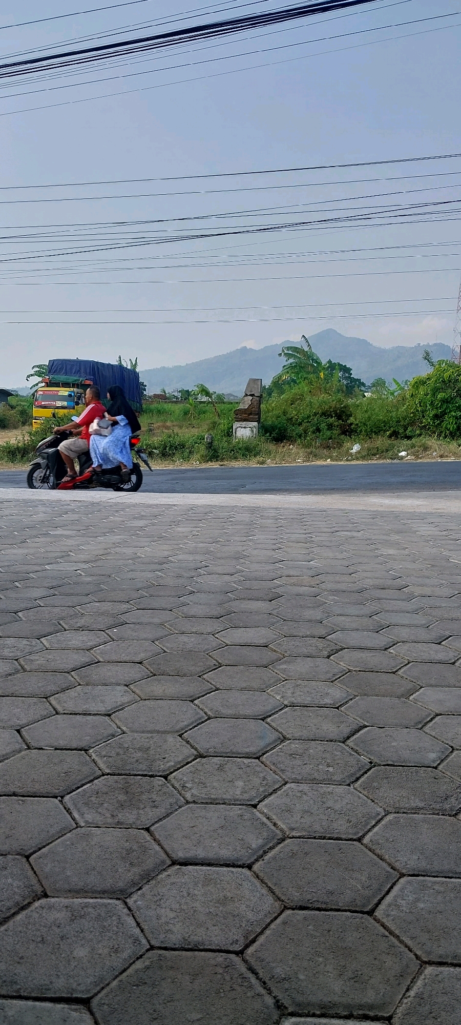 In Keling, Indonesia, a scooter with two individuals is captured in motion on an asphalt road. The driver, a male with a red shirt and dark shorts, is at the controls. A female passenger sits behind him, wearing a blue dress and a black hijab. They are traveling past a roadside scene featuring a yellow truck with a blue tarp, a broken stone structure, and lush green vegetation. In the background, rolling hills are visible under a hazy sky, crisscrossed by overhead power lines. The foreground is dominated by a textured paving of grey hexagonal stones, suggesting a walkway or paved area adjacent to the road.