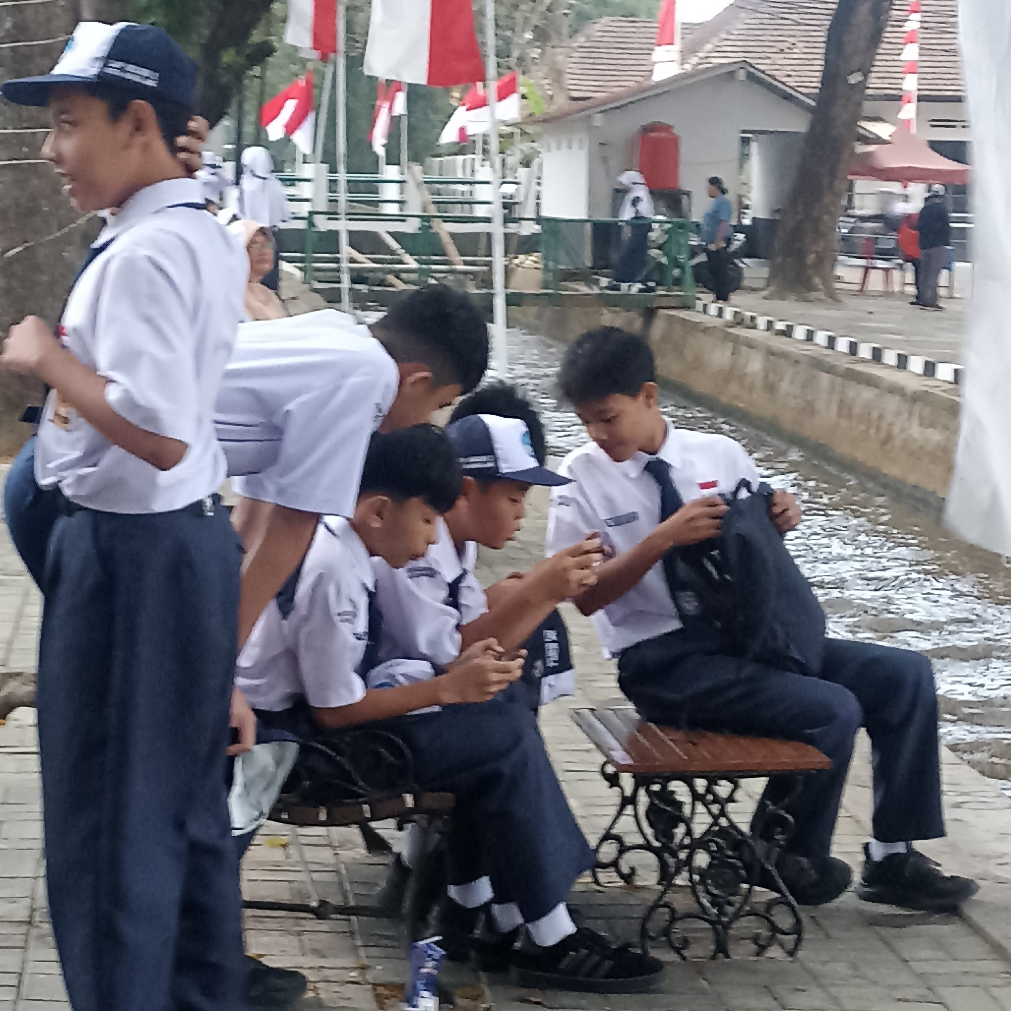 A group of young boys, dressed in school uniforms consisting of white shirts and dark blue pants, are gathered near a body of water. Some are sitting on a bench, while others stand or lean in. Two boys are looking at their mobile phones. In the background, several Indonesian flags are displayed. The scene appears to be an outdoor public area, possibly a park or waterfront, with paved walkways and a building with a tiled roof. The overall impression is one of casual social interaction among students.