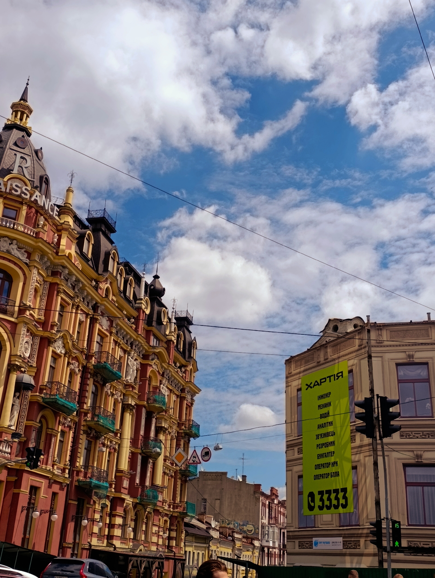 The image captures a view of Khreshchatyk Street in Kyiv, Ukraine, looking upwards towards the sky and the facades of historic buildings. On the left, a grand building, identified by the prominent "RENAISSANCE" lettering on its dome, displays intricate architectural details with red and yellow coloring, ornate balconies, and numerous windows. To the right, a lighter-colored building features a large, bright green banner with Ukrainian text and the number "3333" at the bottom. The banner lists job titles such as "ENGINEER," "MECHANIC," "ANALYST," and "OPERATOR." Traffic lights and overhead wires are visible, indicating an urban setting. The sky is a vibrant blue with scattered white clouds. Several cars are parked at the bottom of the image, and parts of people are visible in the foreground, suggesting the photo was taken from street level. The overall impression is of a sunny day in a bustling city center.