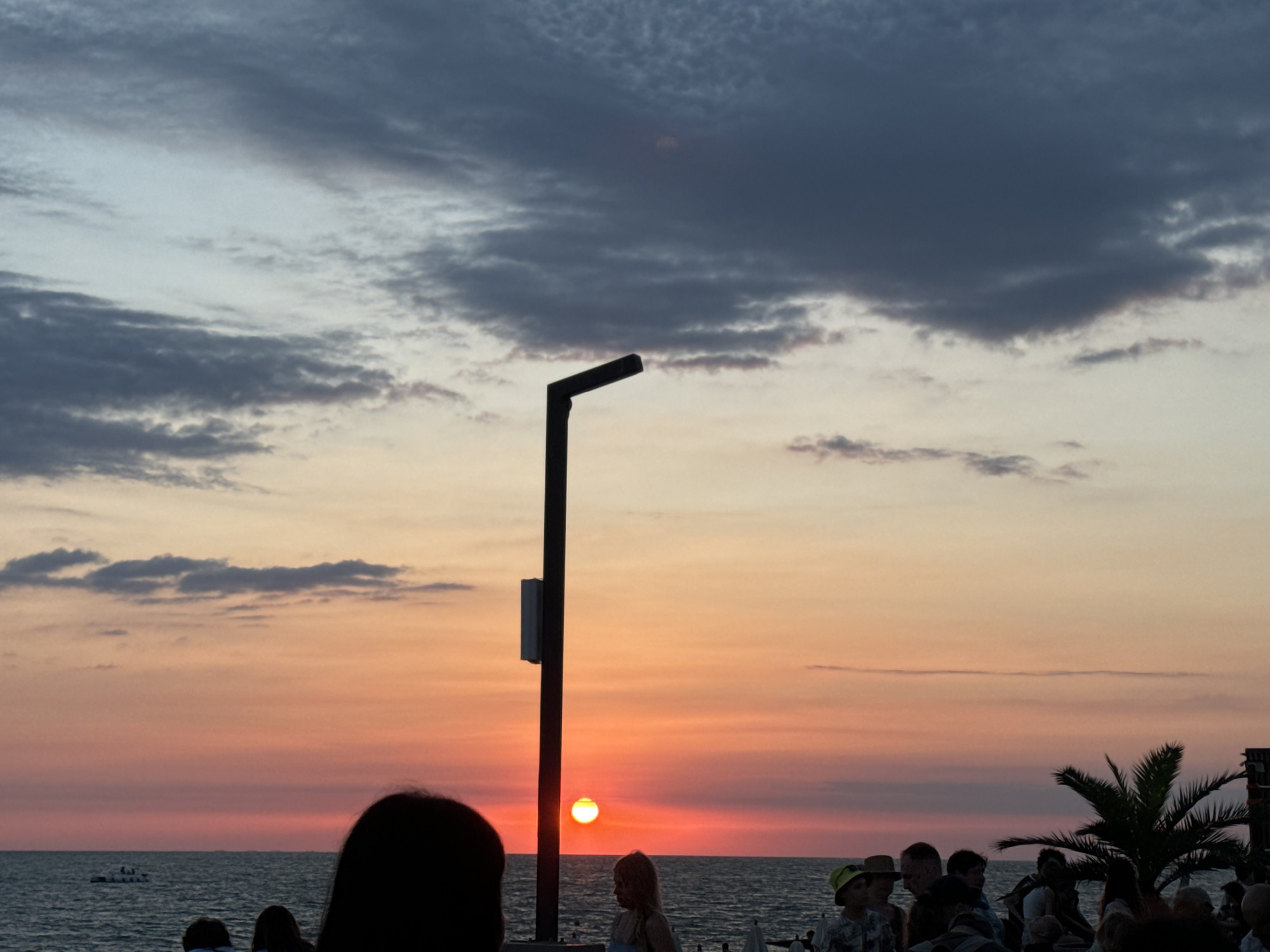 The image captures a sunset over the Black Sea in Sochi, Russia, viewed from a public promenade. The sky is a vibrant canvas of orange, pink, and purple hues, with dramatic dark clouds dominating the upper portion. A solitary lamppost stands prominently in the foreground, silhouetted against the colorful sky. In the middle ground, the sun is setting, appearing as a bright, glowing orb just above the horizon. The sea is a dark expanse of water, with a small boat visible in the distance. A group of people are gathered on the promenade, their forms silhouetted or partially obscured by the fading light. Some individuals are looking out at the sea, while others appear to be engaged in conversation. The overall atmosphere is one of peaceful contemplation and enjoyment of the natural spectacle.