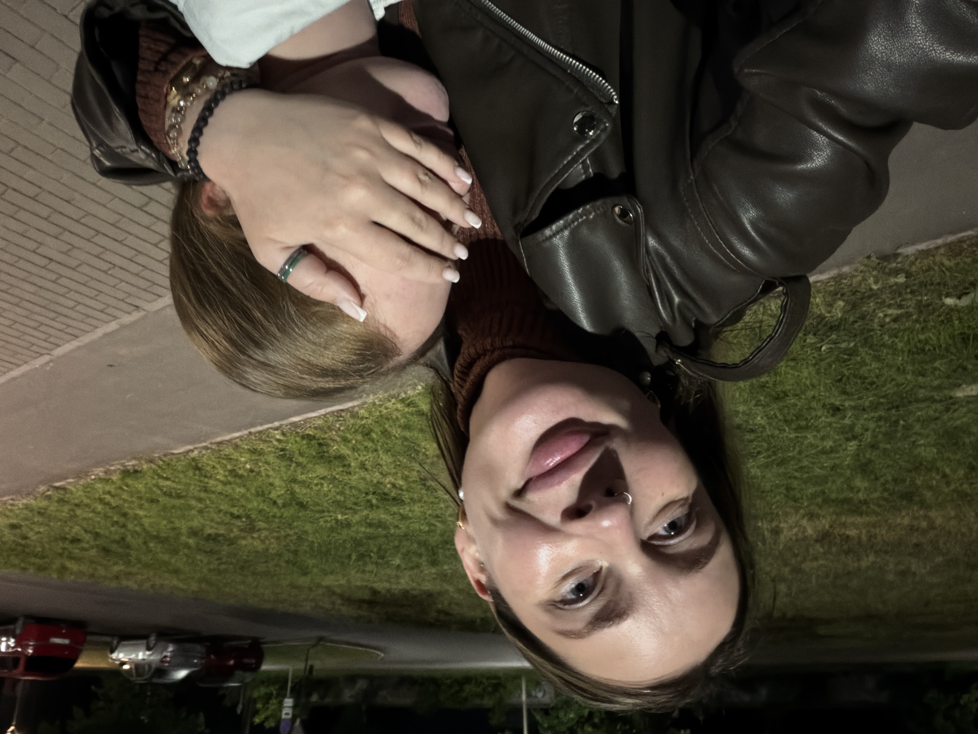 A dimly lit, close-up outdoor portrait captures two individuals in what appears to be a night or late evening setting in Chaikovsky, Russia. The image is oriented as if taken horizontally and then rotated 90 degrees counter-clockwise.
The primary subject is a young woman with light brown hair, positioned on the right side of the frame (when corrected). She is looking directly at the camera with a gentle smile, her lips slightly parted. She has a nose ring on her left nostril and small earrings. Her attire consists of a dark brown leather jacket worn over a brown ribbed turtleneck top. Her right hand is raised, resting on the face of another person beside her, partially obscuring their features. This hand shows long nails with white French tips, a dark green ring on her middle finger, and multiple bracelets on her wrist, including black beaded and gold-toned ones.
The second person, partially visible on the left, has light brown hair and appears to be wearing a white top. Their face is mostly covered by the first woman's hand, suggesting a playful or affectionate interaction.
The background shows an outdoor environment under artificial lighting. There's a paved surface, likely a sidewalk or curb, alongside a patch of green grass. Beyond the grass, a dark road is visible with several parked cars in the distance, including a red, a silver, and a darker colored vehicle. The scene suggests a casual, possibly intimate moment. There is no visible text in the image.