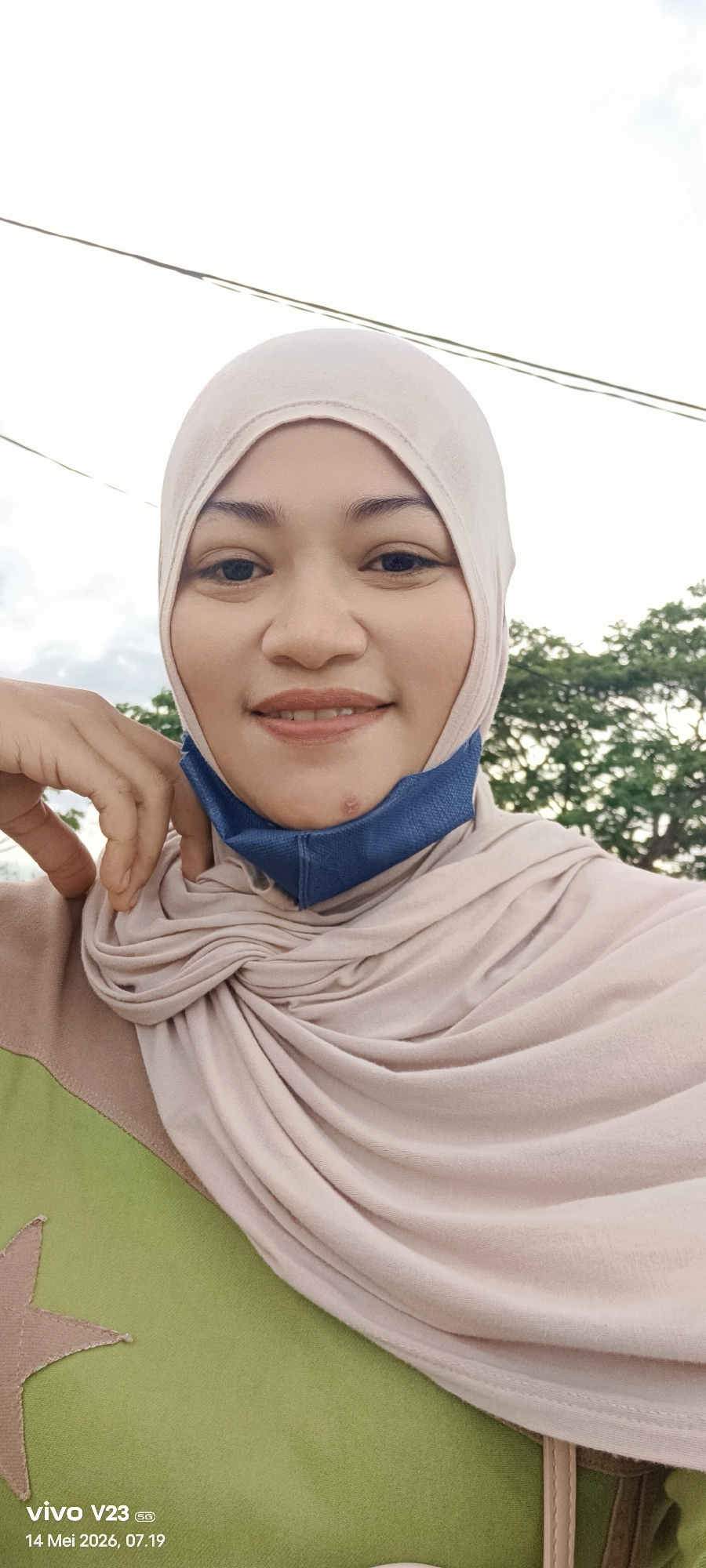 A woman wearing a light beige hijab and a blue face mask is posing for a selfie. She is wearing a shirt with a green section featuring a brown star appliqué, with a beige section next to it. She has one hand resting on her chin. Power lines are visible in the upper left corner of the image, and a tree with green foliage is in the background on the right side. The image has a text overlay in the bottom left corner that reads "vivo V23 5G, 14 Mei 2026, 07.19". The location is Unknown, Unknown.