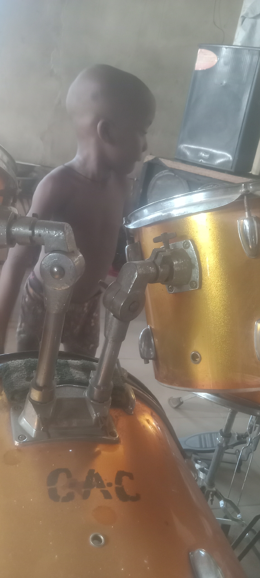 A young, dark-skinned child stands near a drum set indoors in Bende, Nigeria. The child is shirtless and wears patterned shorts. The drum set is primarily gold-colored with silver hardware. The letters "CAC" are stenciled in black on the front drum. A speaker sits behind the drum set. The background is blurry, suggesting a room with light-colored walls.