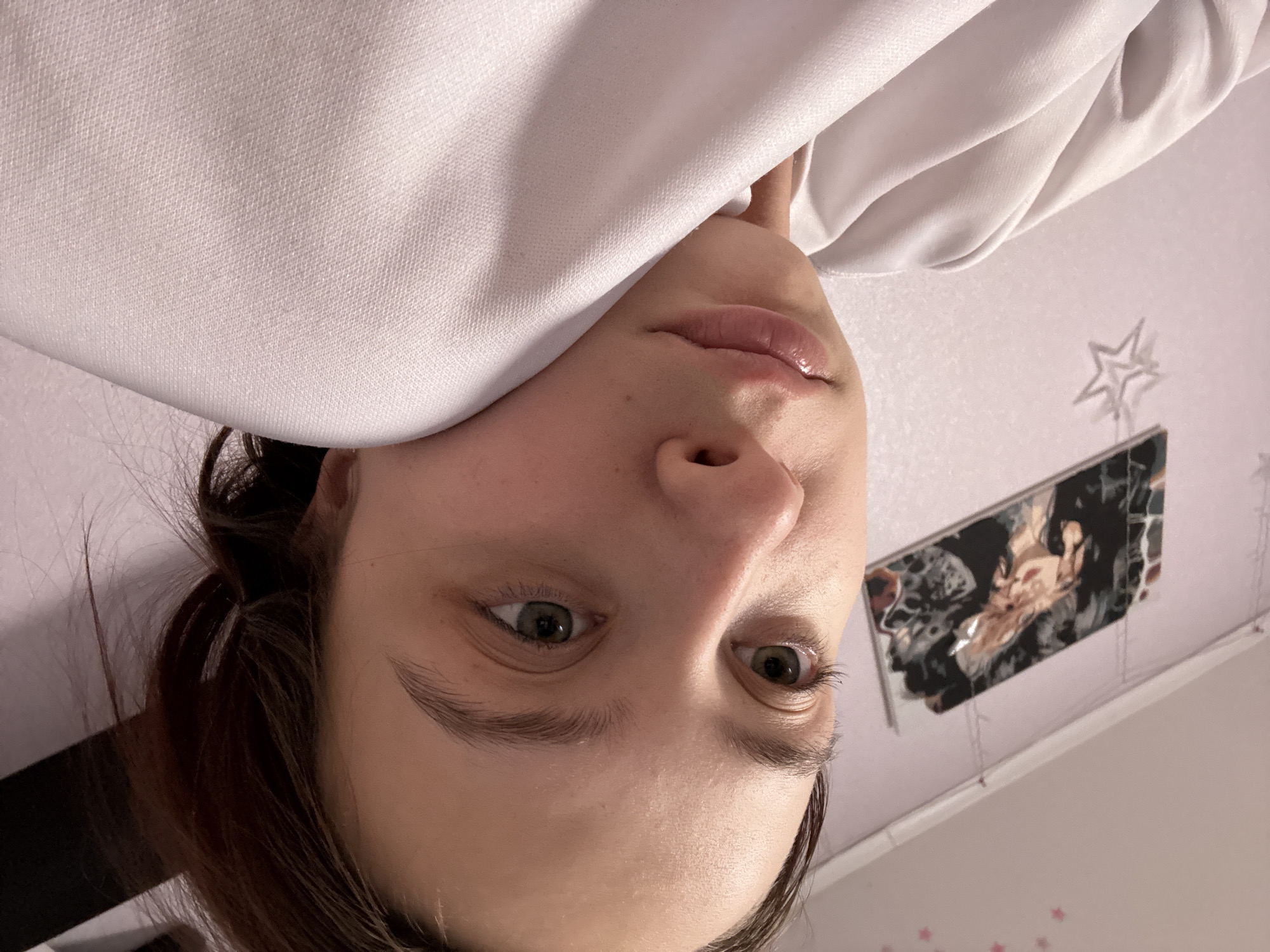 A close-up, inverted shot features a person with light skin, brown hair, and light green eyes. The individual is partially covered with a white garment, possibly a sweatshirt or blanket. The background includes a light pink or white wall adorned with a star-shaped decoration and a rectangular framed image with an abstract black and white pattern. The location cannot be determined from the image, however the user specified the location in the city of Прикамский, Russia.