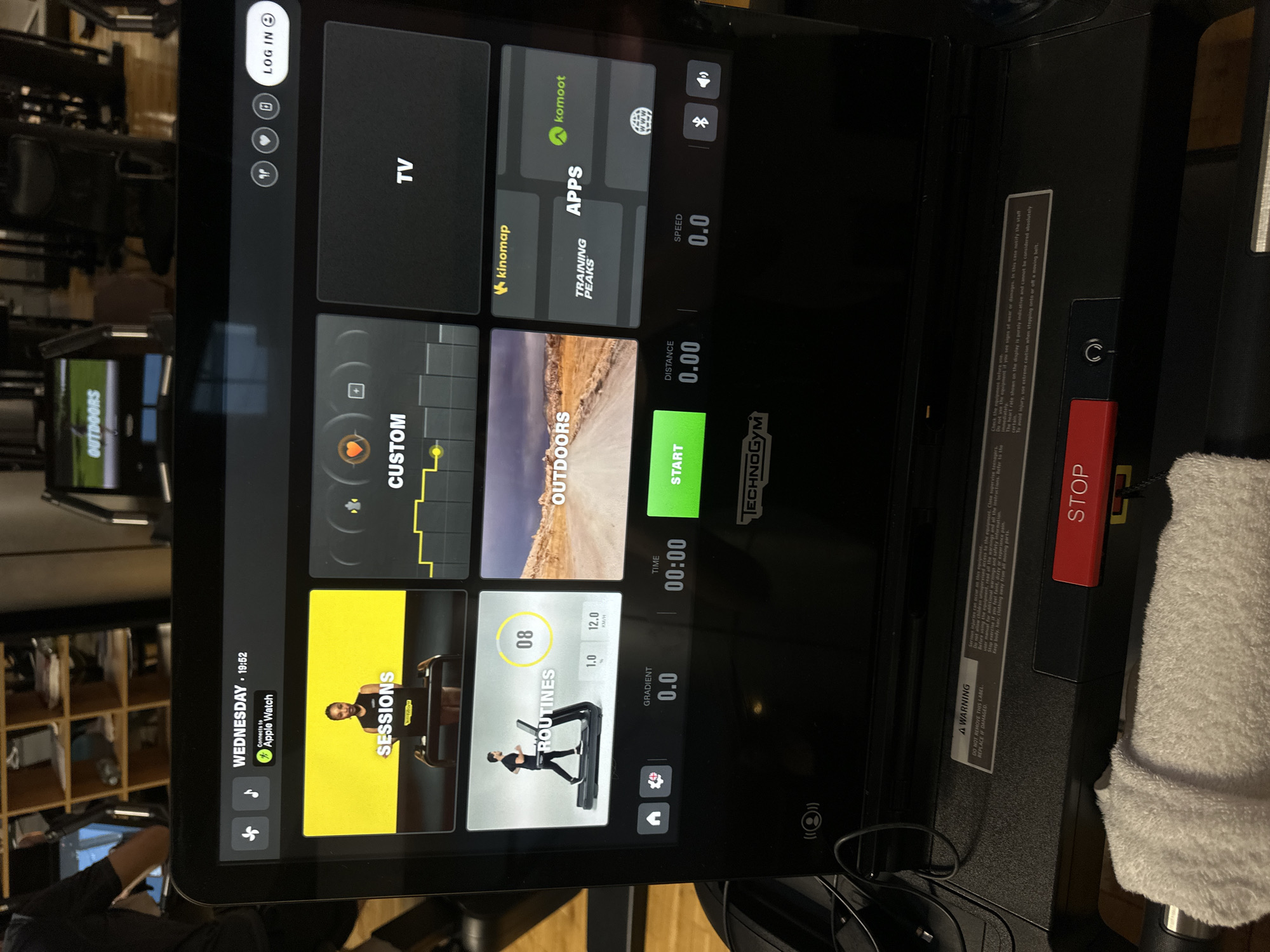 Here is a forensic description of the image:
The image presents a close-up view of a Technogym treadmill interface, likely indoors in a gym setting based on the out-of-focus background showing other exercise equipment. The treadmill display is lit up and shows the main interface screen with options like "Sessions," "Routines," "Outdoors," and "Custom," among others. The screen also shows the current time as 19:52 on Wednesday, and it indicates a connection to an Apple Watch. Several fitness app options are visible, including kinomap and Komoot. The display shows speed, distance, and time as 0.0, 0.00, and 00:00 respectively, indicating the treadmill is currently inactive. A red "STOP" button is positioned on the right side of the treadmill frame, and a white towel is draped nearby. The city is Unknown, Unknown.