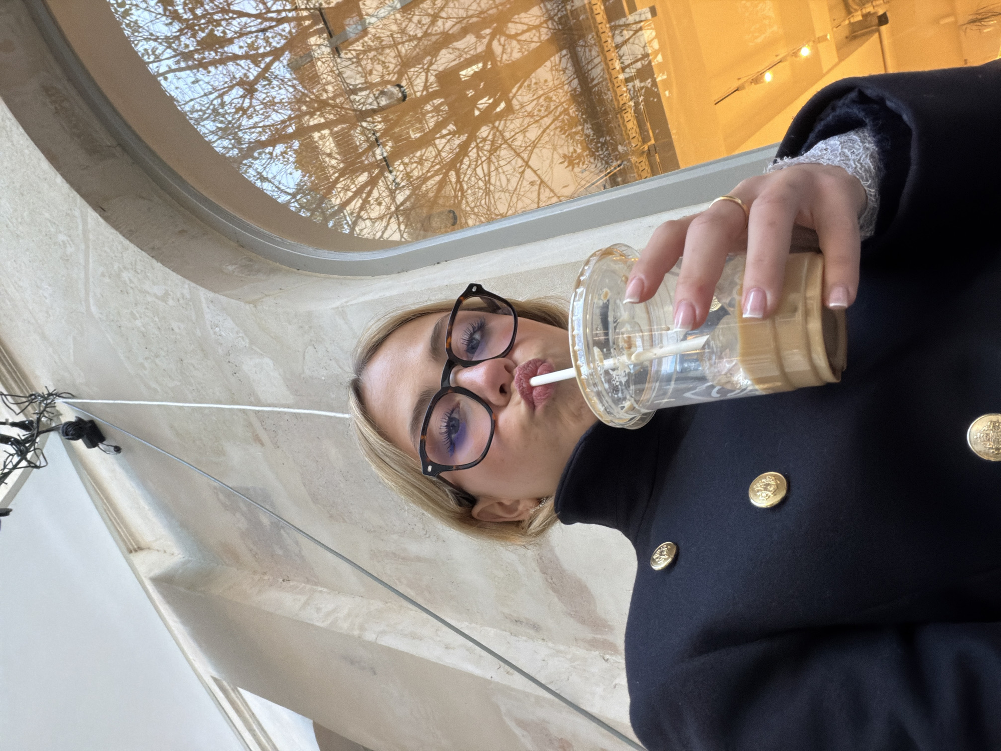 A fair-skinned woman with short blonde hair and tortoise-shell framed glasses is centered, looking directly at the camera while taking a selfie. Her lips are pursed around a white straw, from which she sips a light brown beverage from a clear plastic cup held in her right hand. Her fingernails have a French manicure, and a plain gold ring is on her right ring finger. She wears a dark, high-collared coat or jacket adorned with multiple prominent gold buttons, and a white lace cuff is visible at her right wrist. The background inside features a light-colored, possibly stone or stucco, wall with an arched opening on the right side. Above, wiring and a black fixture are visible against a light ceiling. Through the arched window, reflections of bare tree branches are prominent against a light blue sky, indicating an outdoor urban environment with classic Parisian architecture, identifying the location as Paris, France.