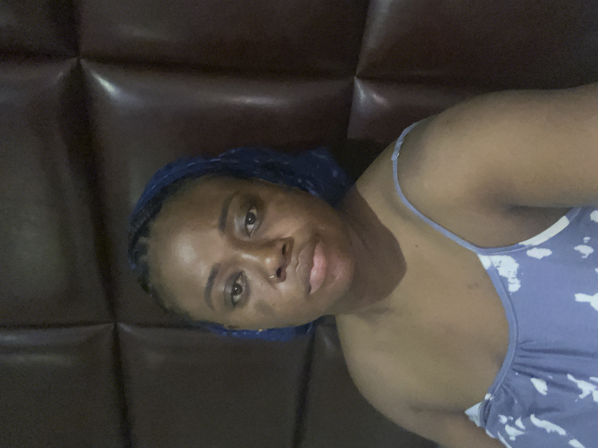 The image is a portrait of a woman with dark skin, captured at an angle, possibly a selfie. She is wearing a blue durag or head covering that reveals parts of her braided hair underneath, and a light blue sleeveless top adorned with white, cloud-like patterns. A small ring piercing is visible in her right nostril. Her eyes are open and directed towards the camera, with a neutral to slightly relaxed expression on her face. The background is a dark brown, padded or quilted surface, likely part of a headboard or couch, indicating an indoor setting. The lighting is soft, suggesting artificial illumination rather than strong natural light. No other individuals, prominent objects, or text are visible in the frame. The image does not contain visual cues to specifically identify the time of day, weather, or the provided location context of Abuja, Nigeria.