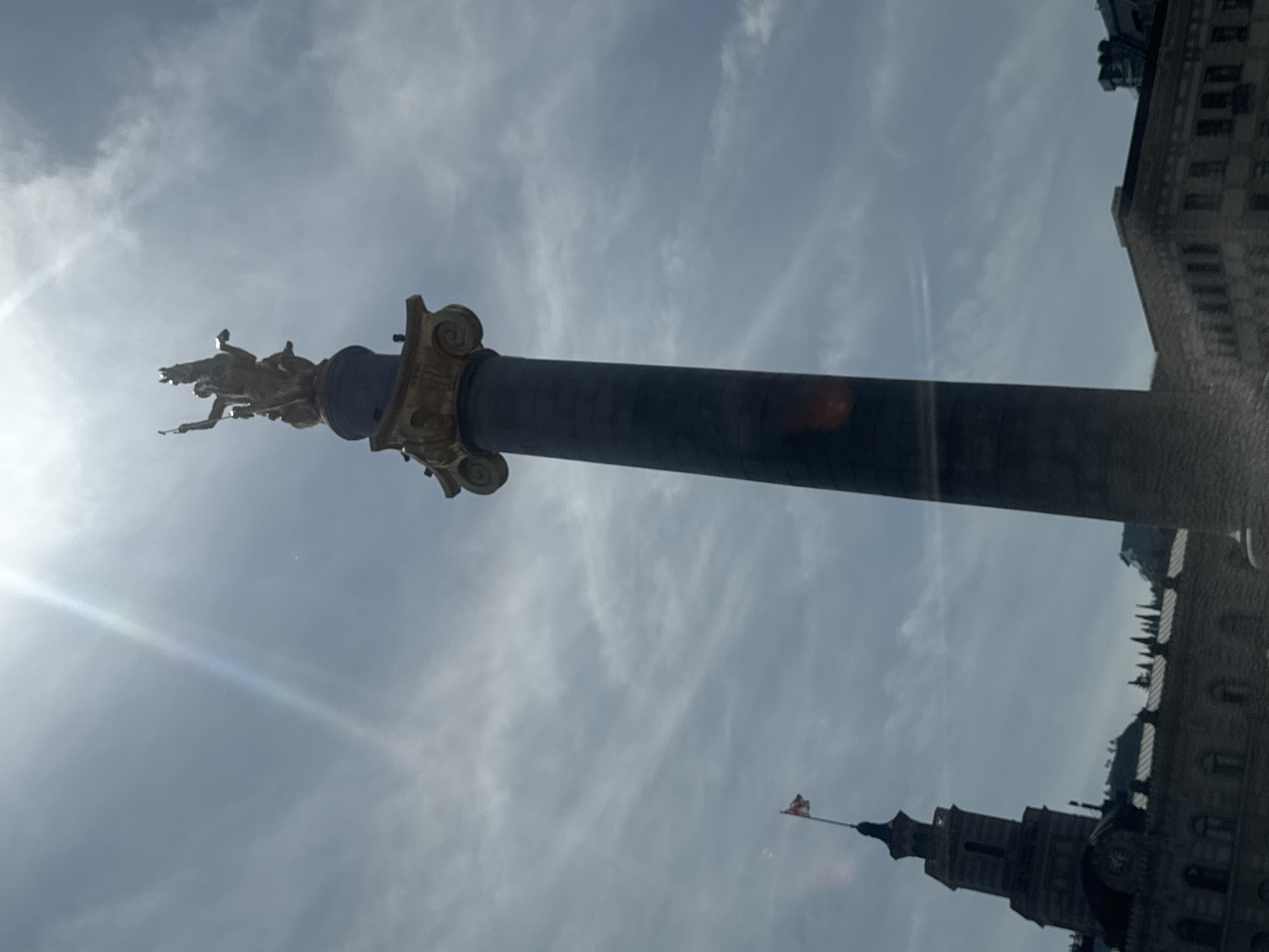 The image depicts the Saint George Statue on the Freedom Monument in Tbilisi, Georgia, shot from a low angle, looking upwards. The monument is a tall column topped with a golden statue of Saint George slaying the dragon. The background shows a cloudy sky, and parts of the surrounding buildings. The flag of Georgia is visible on a building near the bottom. The lens flare from the sun is visible on the left side of the image.