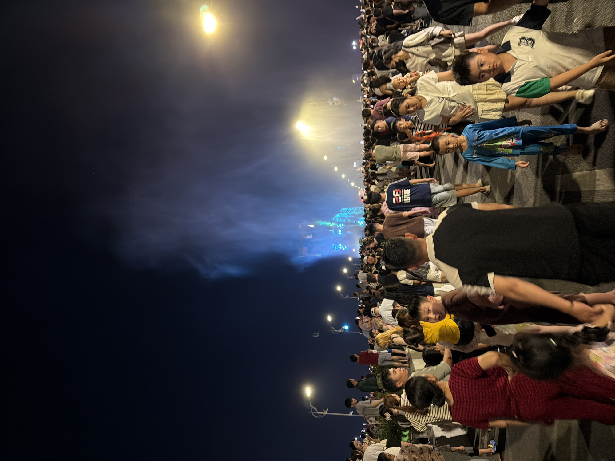 A nighttime scene in Đà Nẵng, Vietnam, depicts a large crowd of people, including adults and children, gathered along a paved area resembling a wide sidewalk or street. Many are sitting or lying down on mats or blankets. Streetlights illuminate the area. In the background, there is a haze or smoke, potentially from a nearby event or activity, possibly fireworks or a light show. The sky is dark. The people appear to be relaxed and watching something in the direction of the haze.