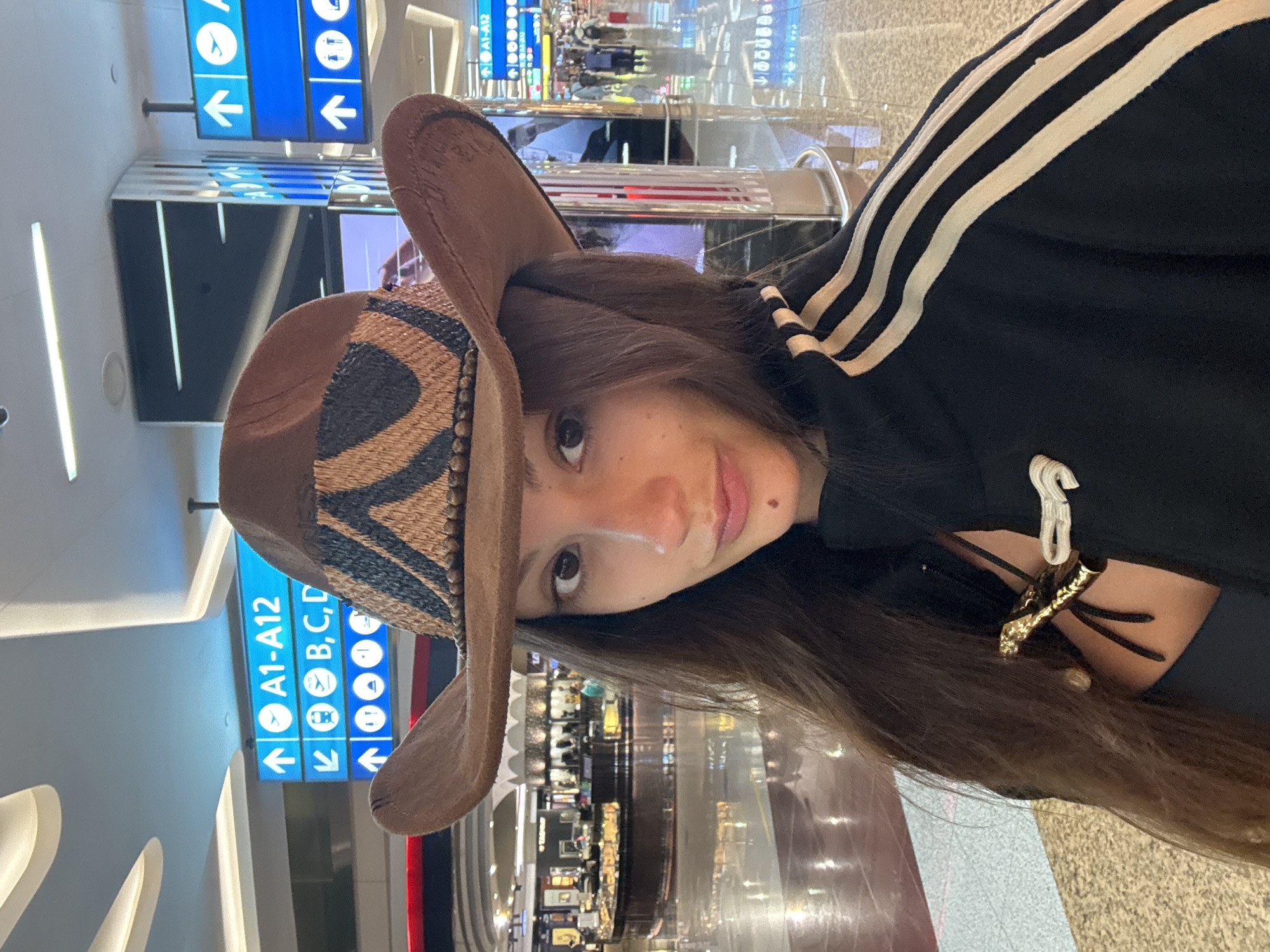A medium shot shows a young Caucasian woman wearing a brown cowboy hat and a black jacket with white stripes. She is standing in an airport terminal, likely Dubai International Airport (DXB), based on the signage. The background shows typical airport features: a ceiling with modern lighting, blue directional signs indicating gates and services, shops with display windows, and other travelers walking. The woman is looking towards the camera with a neutral expression. Her hat has a decorative band with a geometric pattern. The jacket appears to be a tracksuit jacket. There is a decorative clasp on the jacket.