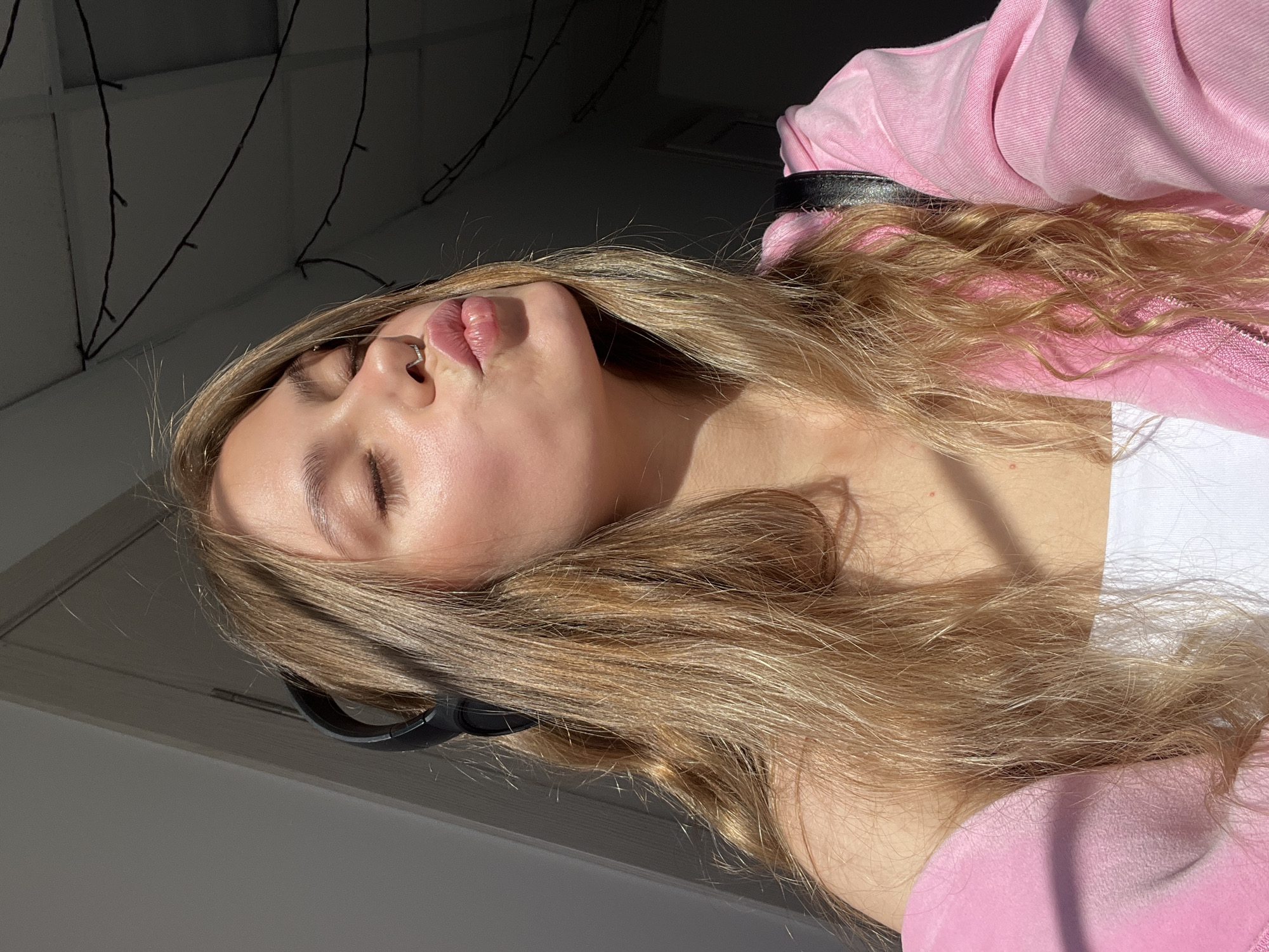 The image shows a young woman with long, blonde hair and closed eyes, puckering her lips. She is wearing headphones and a pink jacket over a white top. She has a septum piercing. The lighting suggests it is daytime, with sunlight casting shadows on her face and hair. The background is dimly lit and appears to be indoors, with what looks like strings of lights visible on the ceiling. There are no identifiable landmarks or contextual clues that would place the image specifically in the city of Фоки, Russia.