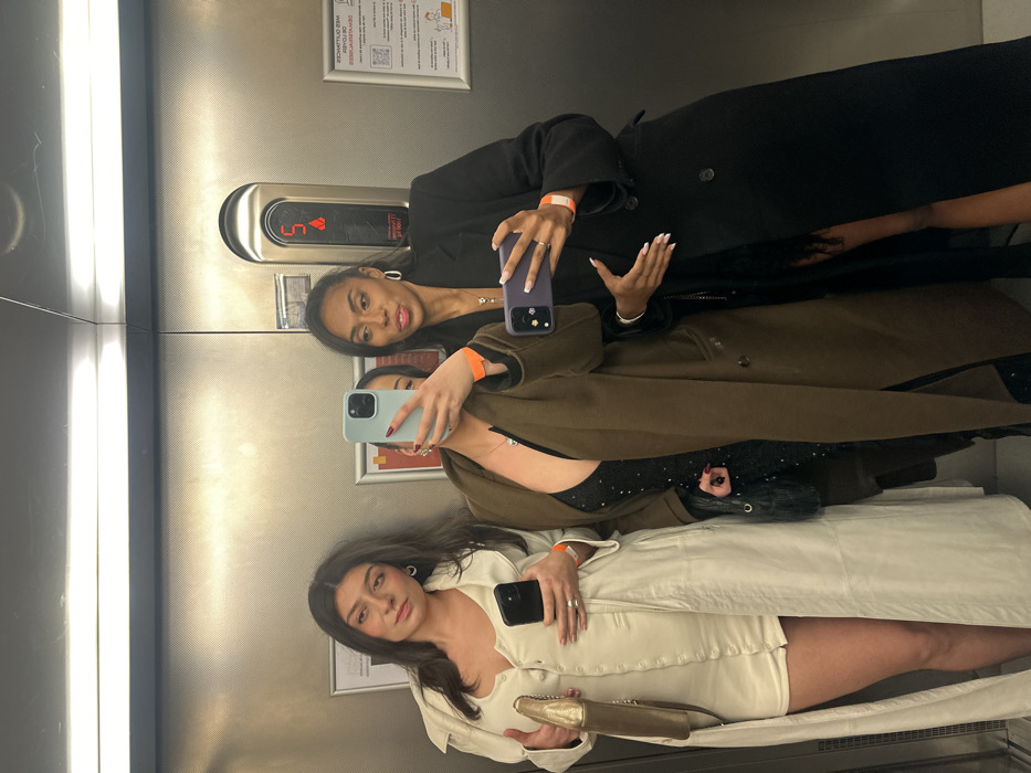 Three women pose for a selfie in an elevator. The first woman wears a black coat, silver hoop earrings, and holds a purple phone. The second woman wears a brown coat, orange wristband, and green phone. A black purse hangs off her shoulder. The third woman, in the reflection at the bottom, wears a cream-colored coat and dress, holding a gold clutch and a black phone. All three have matching orange wristbands. The elevator has a steel interior and a lit-up floor indicator.