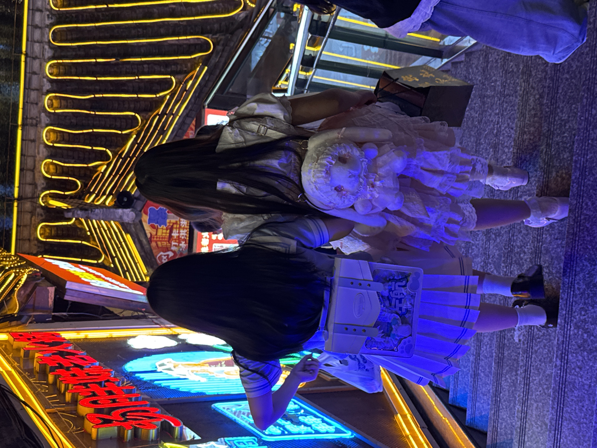 Two young women are standing next to each other on a set of outdoor stone stairs, illuminated by numerous neon signs. The woman on the left wears a pale white lolita dress and holds a matching stuffed toy, while the woman on the right wears a striped top and a short, pleated skirt, carrying a white bag with graphics on it. They both have long, dark hair that hangs down their backs. The background features dense, vibrant neon signs in various colors, including red, blue, and yellow, with Chinese characters. The location is in Yuzhong District, China, judging by the style of the signs and the urban architecture.