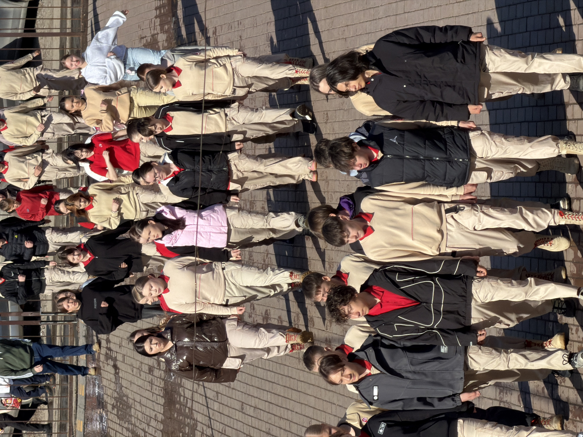 A large group of people lies in rows on a paved surface. The paved surface consists of rectangular bricks, creating a striped pattern. The people are dressed in a variety of clothing, including light-colored jumpsuits, dark jackets, red shirts, and a shiny brown jacket. Some individuals are wearing shoes, while others appear to be barefoot. Some of the people are wearing white clothes with a bit of blue color. The location appears to be outdoors. The city of Izhevsk, Russia is specified as the location.