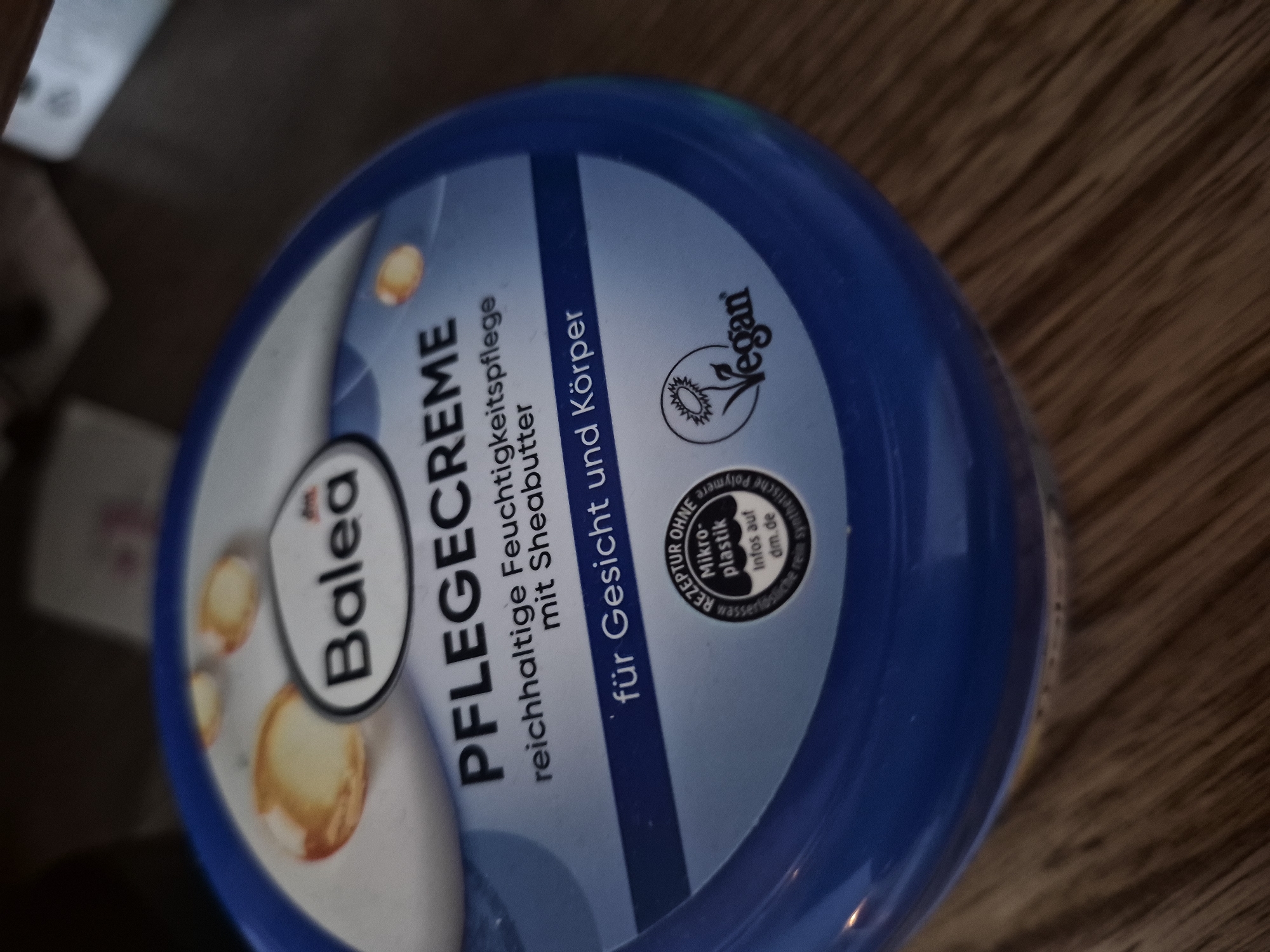 A blue plastic jar of Balea Pflegecreme with shea butter is positioned on a wooden surface. The jar has text in German and a circular emblem indicating "Rezeptur ohne Mikro-Plastik" (Recipe without micro-plastics) and another emblem showing a flower with the word "Vegan" next to it. The jar is partially illuminated, with some parts in shadow.