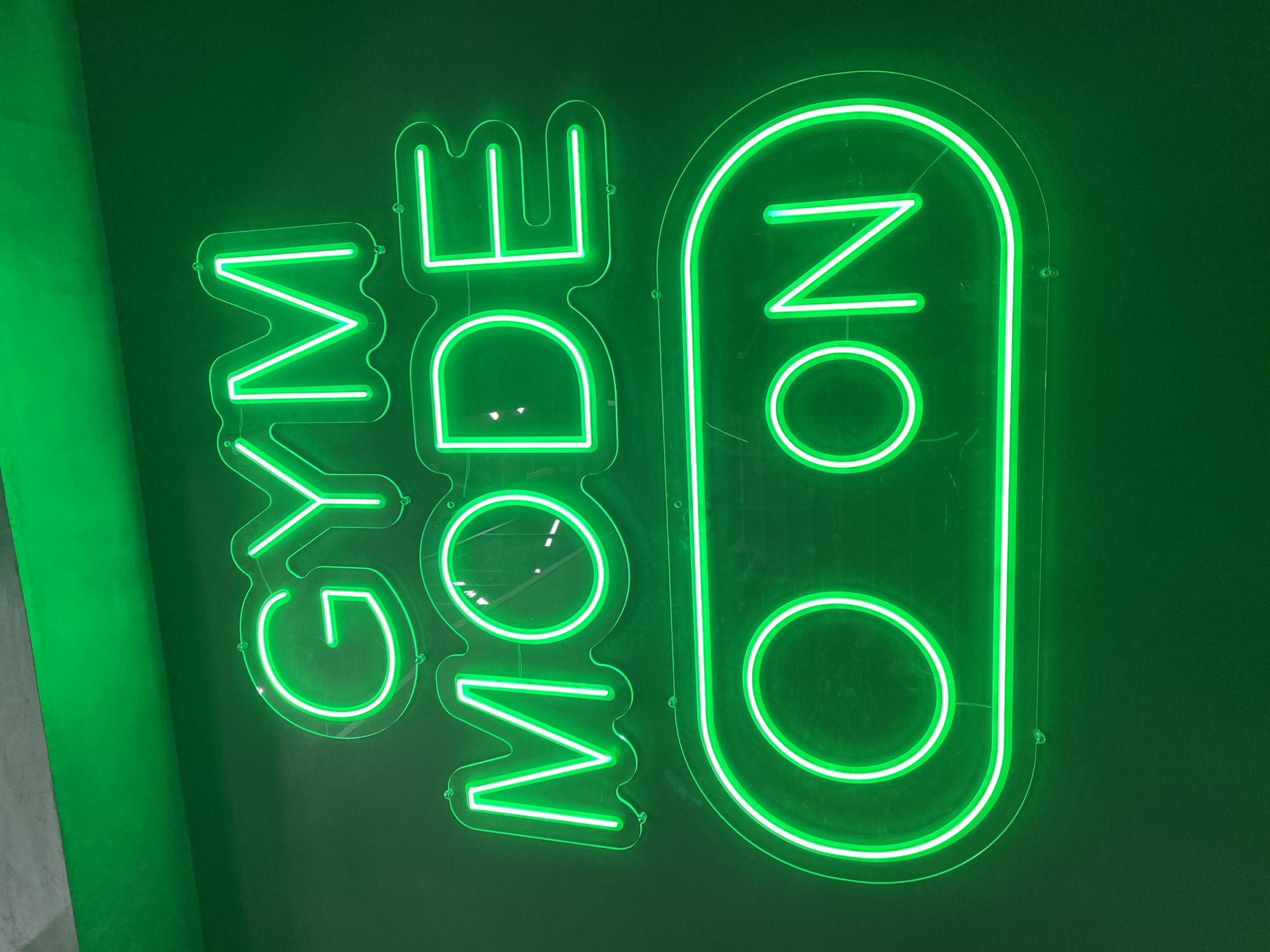 A neon green sign displays the words "GYM MODE ON" on a dark green wall. The words "GYM MODE" are stacked vertically, followed by the word "ON" inside a rounded rectangle, resembling a power switch. The location is unknown, and no details suggest it is in Plovdiv, Bulgaria.