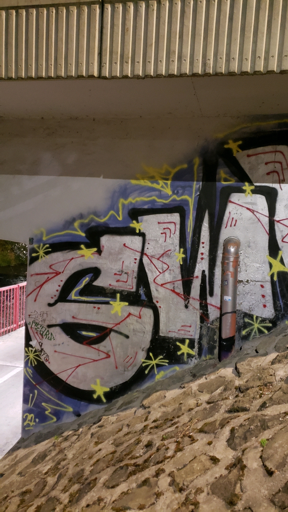 The image shows a section of graffiti on a concrete wall beneath an elevated structure, likely a bridge or overpass. The graffiti is colorful, featuring large, stylized letters in silver and black outlines, with accents of red and yellow. Small yellow star-like shapes are scattered around the lettering. A metal pipe, orange and silver, is visible on the right side of the graffiti. To the left of the graffiti, a pink metal railing can be seen, suggesting a pathway or walkway nearby. The wall itself appears to be made of concrete, with some texture and possibly signs of age or wear. The foreground is dominated by a rough, stone-covered embankment. The overall scene appears to be outdoors, under artificial lighting, and likely in an urban or semi-urban environment. There is no indication of specific people, actions, or interactions. The location is identified as being in the city of Prešov, Slovakia.
