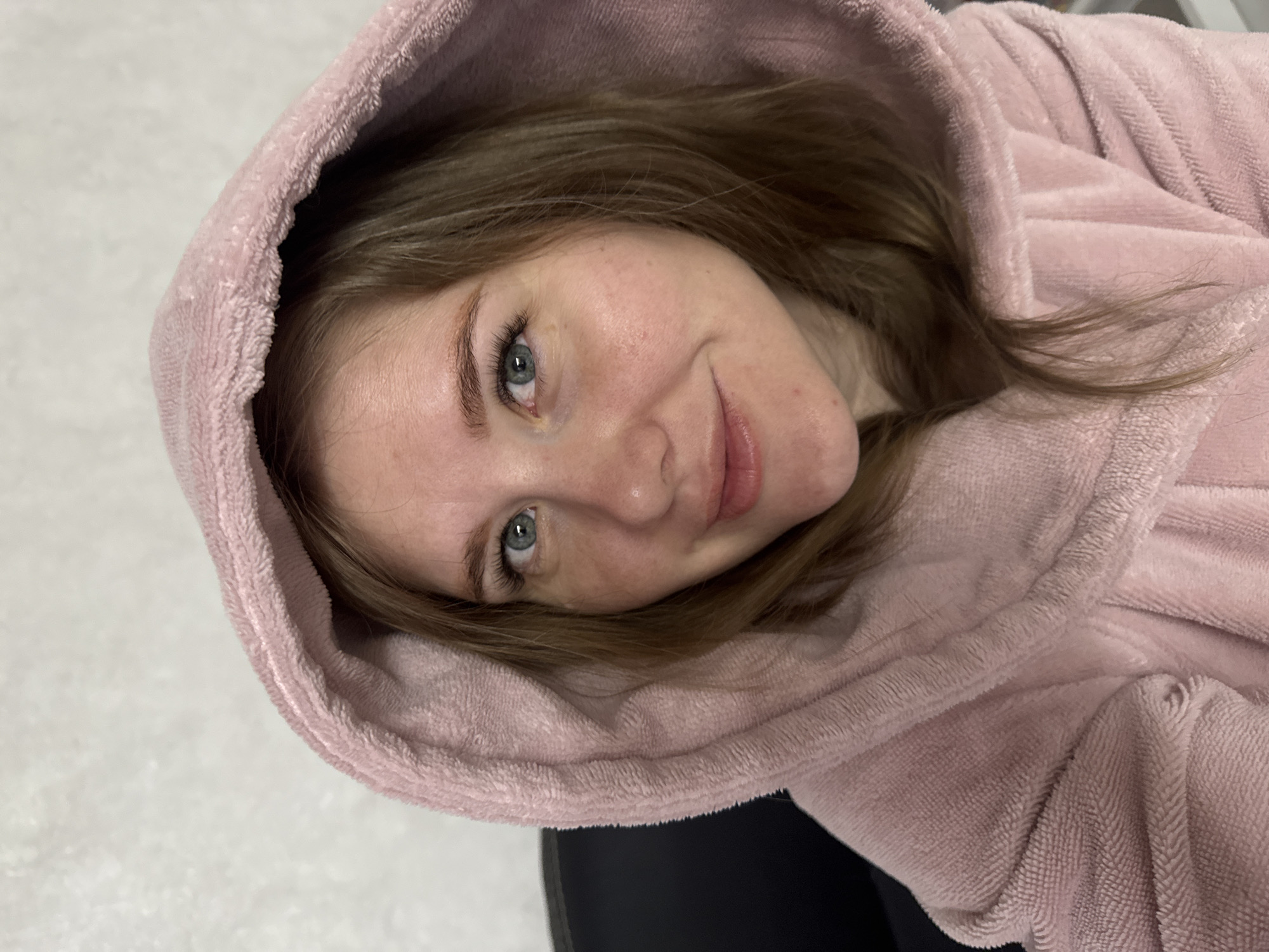 The primary subject of the image is a young woman, captured in a close-up portrait. She is wearing a plush, soft pink hooded garment, possibly a robe or a hooded blanket, made of a towel-like or fleece material, with visible folds and texture. A hint of dark clothing is visible below her right shoulder.
The scene is indoors, presenting a relaxed and casual atmosphere. The background on the left is a light, textured surface, while the right side of the background is darker and less defined.
The woman is posing for the camera, with her head tilted, and she is looking directly into the lens. She has a gentle, subtle smile on her face, conveying a sense of calm or contentment. Her eyes are blue-green, and her eyelashes appear long and dark. Her hair is light brown or auburn, flowing out from under the hood and framing her face. She has fair skin with visible freckles across her nose and cheeks. There are no specific visual cues within the image to denote the time of day, weather, or any precise location within Gornyi (Dvurechenskoe S/P), Russia, beyond it being an indoor setting. No visible text is present in the media file.