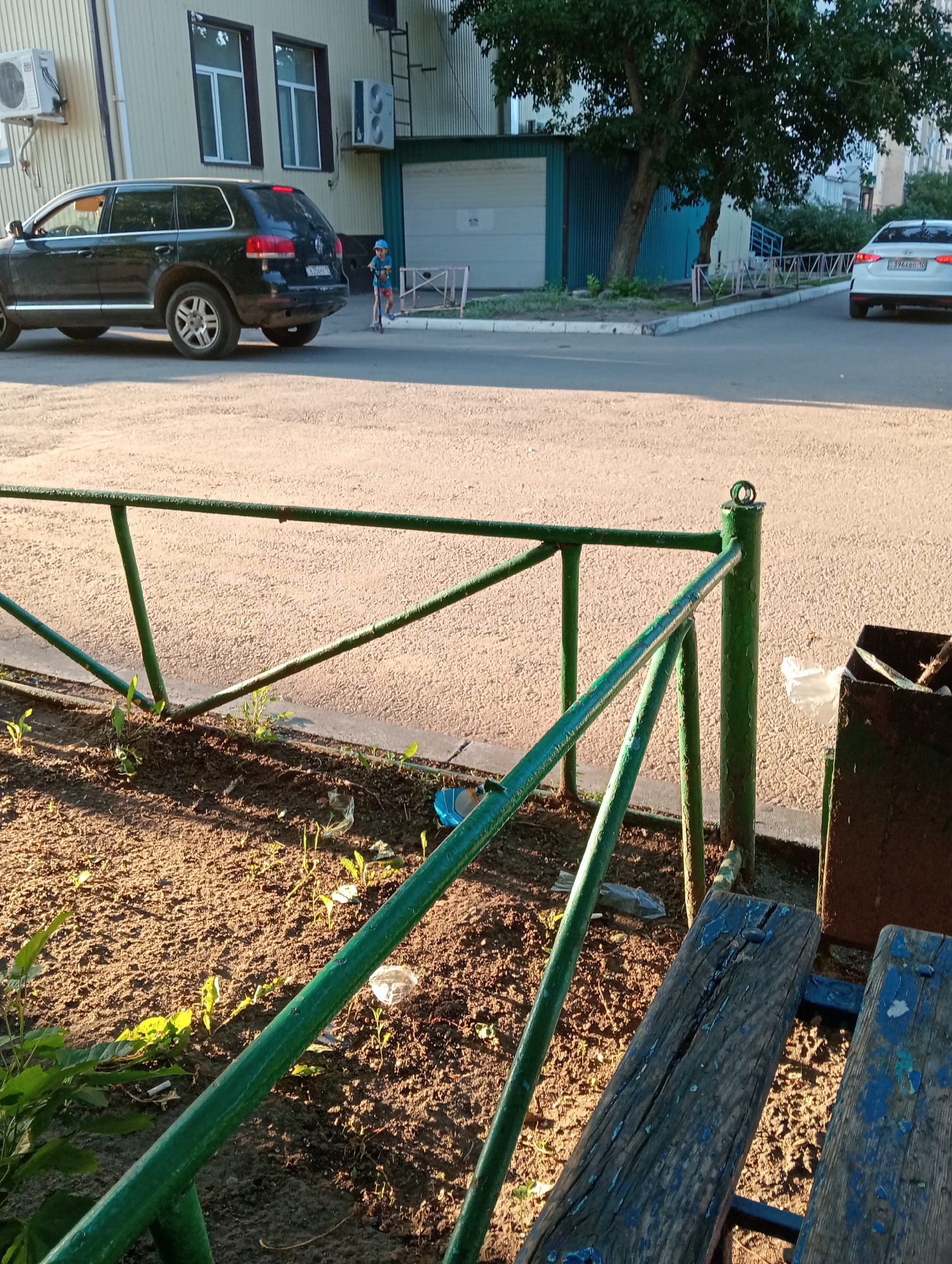 In a suburban setting, an SUV is parked on the side of a paved road, adjacent to a building with light-colored walls and dark-trimmed windows. A child on a scooter is near the SUV, facing away from the camera. A white car is visible in the background, further down the road. The foreground features a dirt patch with sparse vegetation and a green metal fence. Two weathered wooden benches are also present. A dark metal trash receptacle stands next to the fence. The scene is illuminated by the warm light of what appears to be late afternoon or early evening sun, casting long shadows.