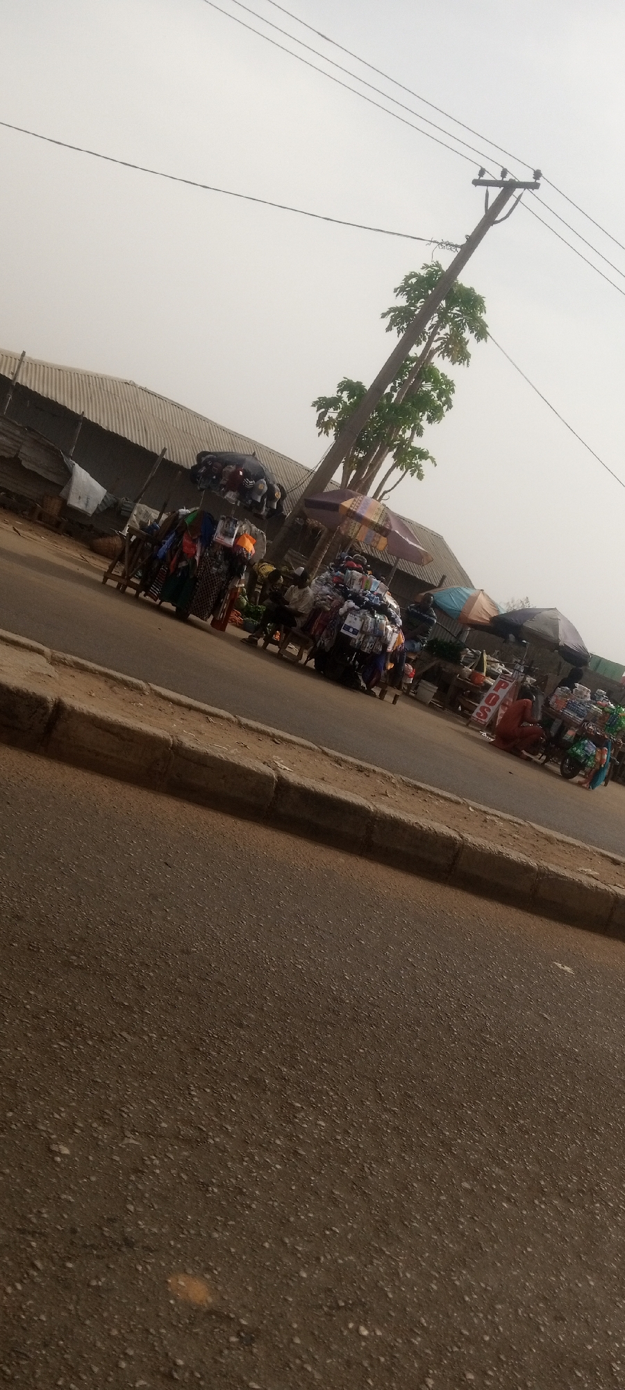 This media file captures a street scene in Sabon Tasha, Nigeria, depicting a bustling roadside market under a hazy sky. The foreground features a dusty asphalt road with a concrete curb running along its edge.
In the midground, numerous makeshift market stalls are set up along the roadside, brimming with a variety of goods. These appear to include clothing, textiles, and general merchandise, piled high for display. Several large umbrellas, some colorful and striped, provide shade over the stalls and vendors. A tall, leafy tree, possibly a papaya tree, stands behind some stalls, adjacent to a wooden utility pole with multiple overhead power lines stretching across the upper portion of the frame.
Several people are visible engaging in commerce. One individual, dressed in a reddish-brown garment, is crouched near a stall that features a white sign with red lettering clearly stating "P.O.S." (Point of Sale), indicating a financial transaction service. Other vendors are present behind their respective stalls.
The background shows buildings with corrugated iron roofs, typical of local structures in the region. The sky is a uniform, light grey, suggesting either overcast weather or a dusty atmospheric haze, common in certain seasons. The lighting is bright but diffused, casting soft shadows. The overall scene portrays a vibrant, everyday local market activity.