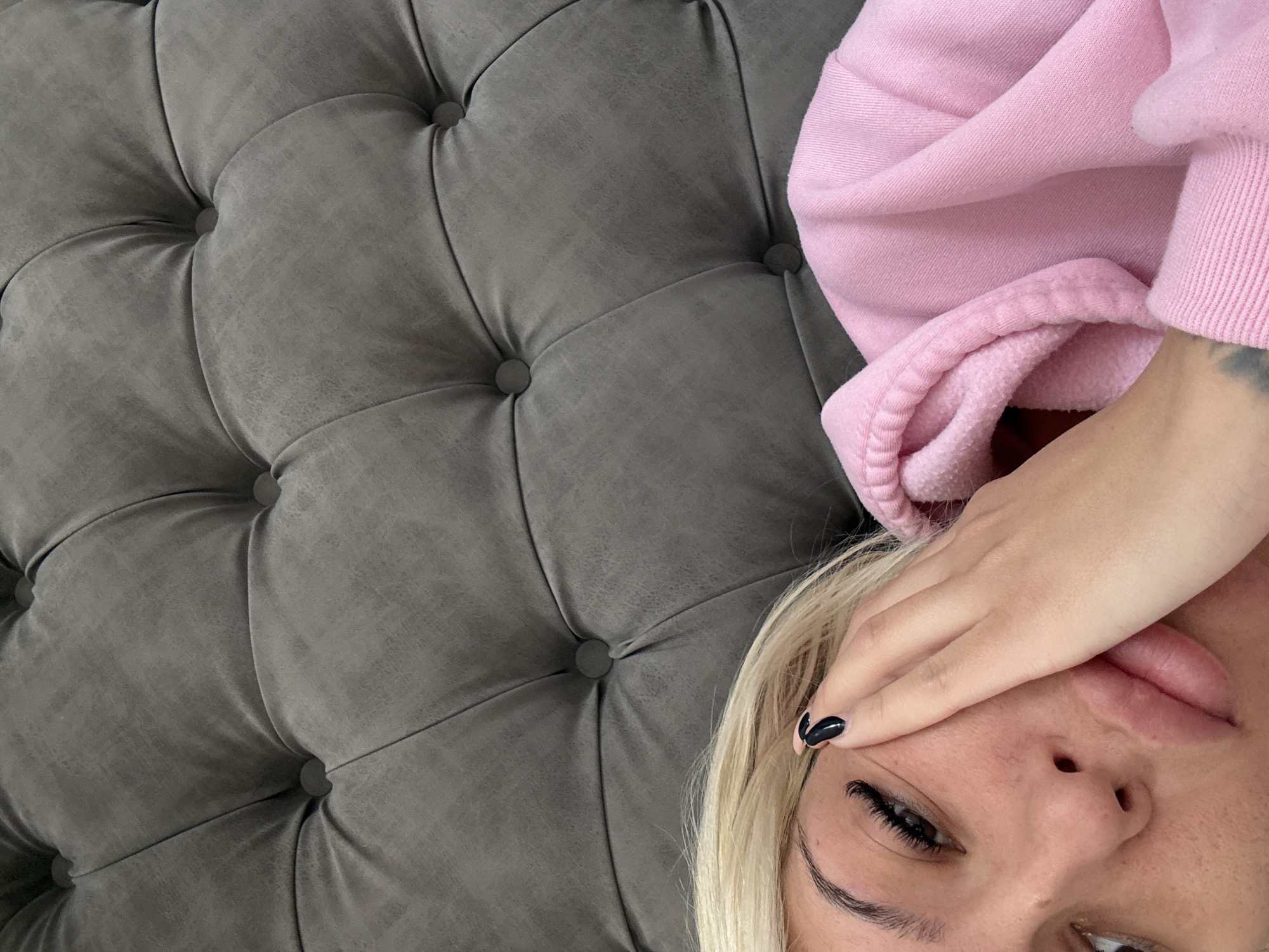 A selfie-style, close-up shot shows a person lying on a gray, tufted surface. The person is wearing a pink sweatshirt, and part of their face is visible, including the nose, mouth, eye, and blonde hair. Their hand covers part of their face, with black nail polish visible. The background is dominated by the tufted gray surface. The location is Unknown, Unknown.