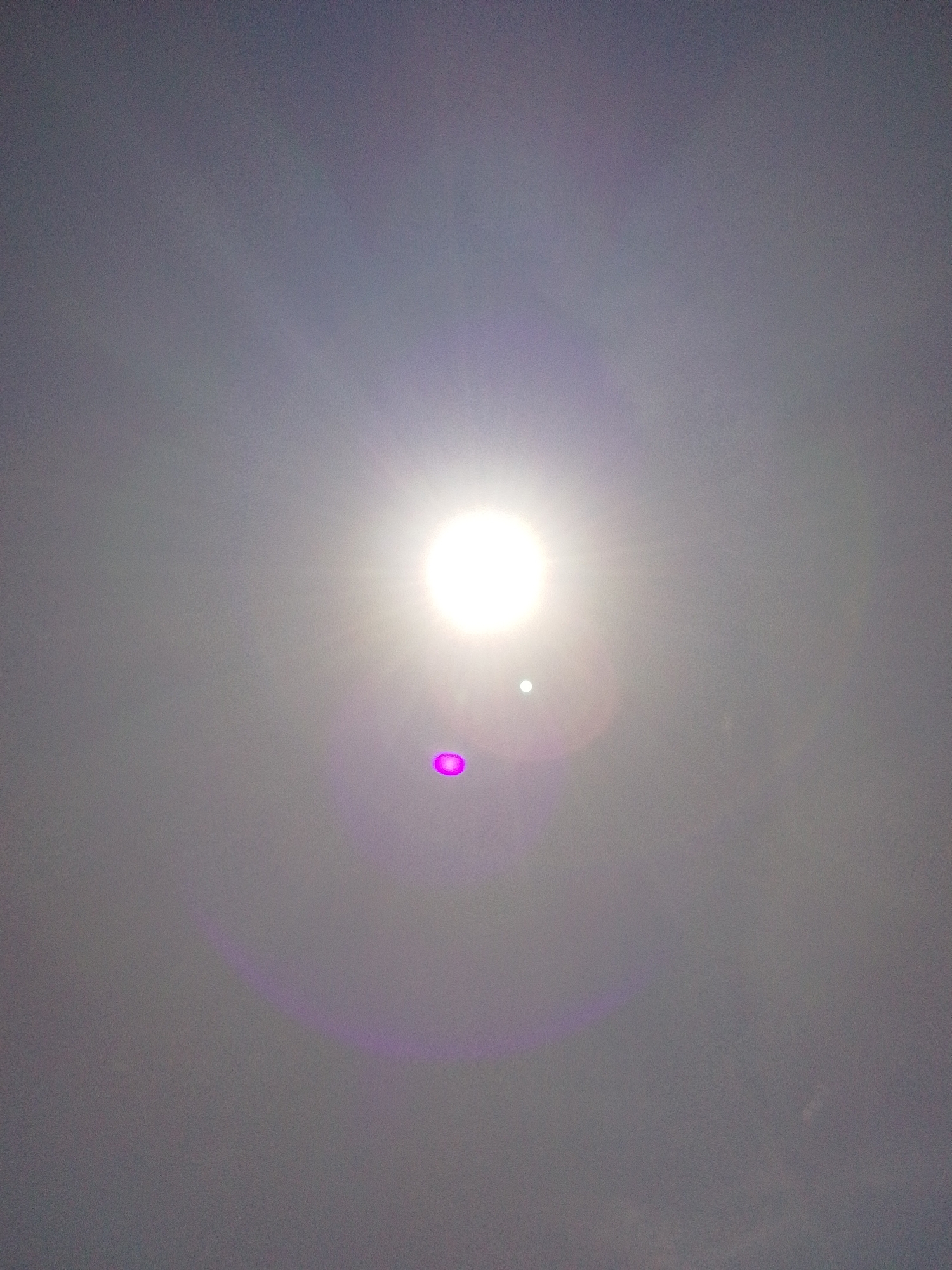 The image is a photograph of the sky, likely taken during daylight hours. The sun is visible as a bright, blown-out disc in the upper center of the frame, emitting strong rays that create a lens flare effect. There are also two distinct, circular lens flares present: a smaller, white-ish one below the sun and a larger, vibrant magenta one further down and to the left. The sky itself appears hazy or overcast, with a muted greyish-purple hue. The overall impression is of a bright, sunlit sky with optical artifacts from the camera lens. No people, objects, actions, or interactions are discernible in the image. The location in Serti, Nigeria, cannot be determined from the visual content of the photograph.