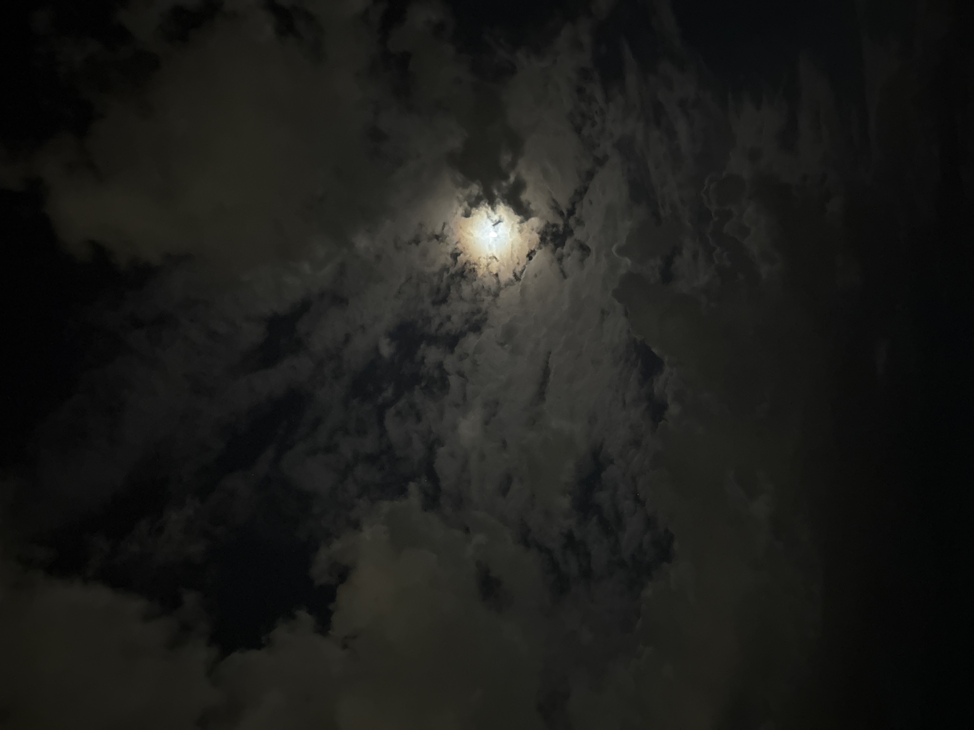 The image depicts a night sky with clouds partially obscuring the moon. The moon is visible as a bright, circular shape in the center of the frame, surrounded by varying densities of clouds. The overall scene appears dark due to the night setting. There is no visible information indicating the location is in Plovdiv, Bulgaria, and the image lacks details about people, subjects, objects, or actions.