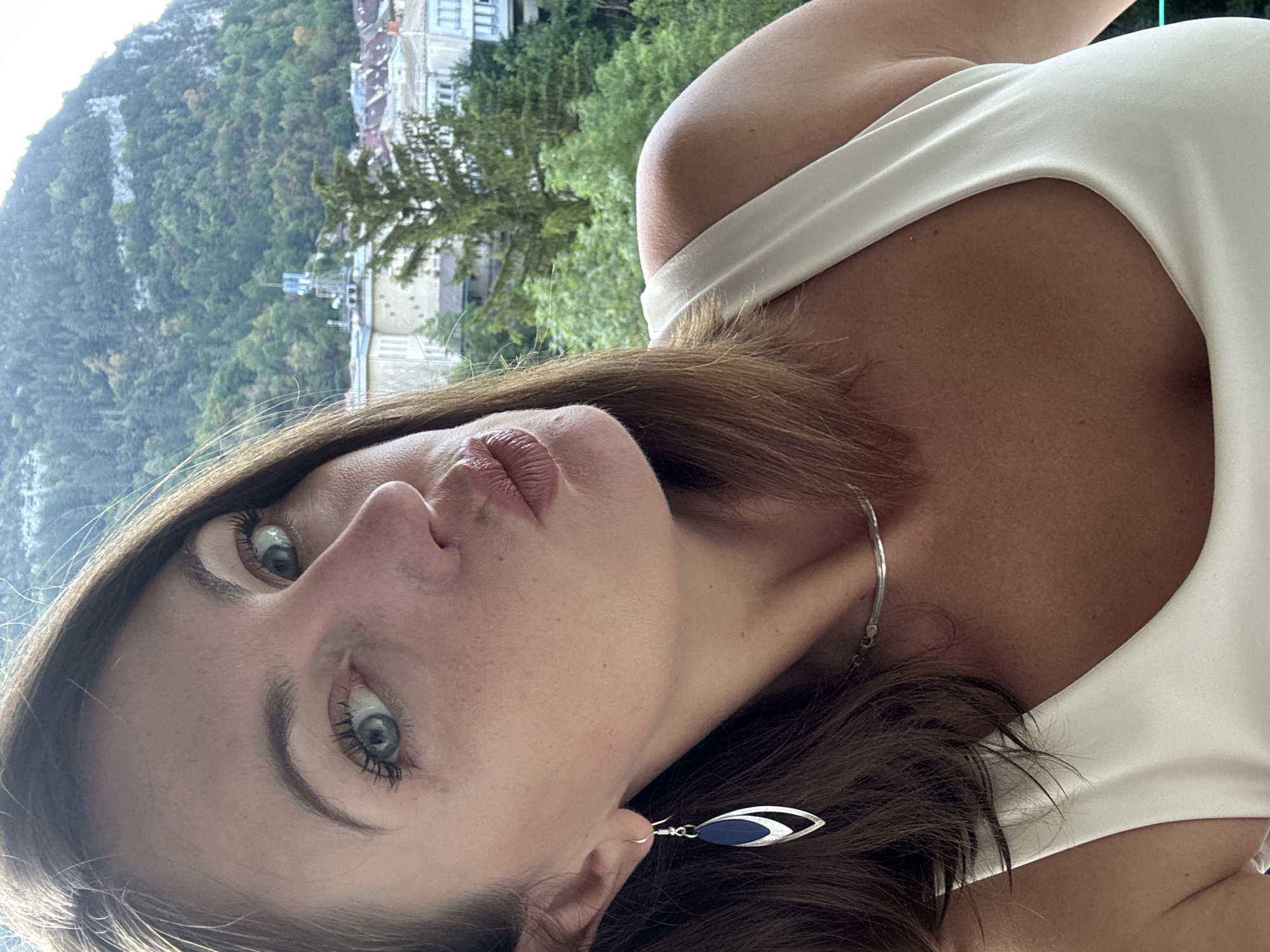 A close-up, selfie-style shot features a woman with brown hair and blue-green eyes, wearing a light-colored tank top, a silver necklace, and an earring. Her lips are pursed as if for a kiss. In the background, a mountain covered in trees and a town with buildings and trees are visible. The scene appears to be in Saint-Claude, France.