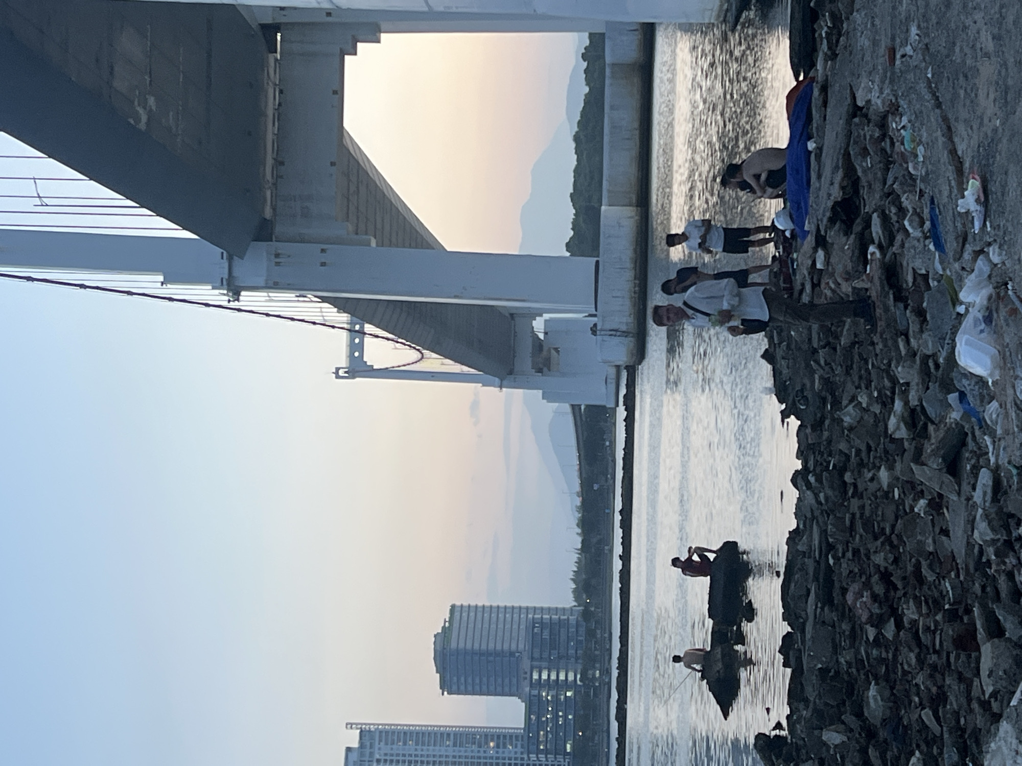 The image shows a scene near the Dragon Bridge in Đà Nẵng, Vietnam, at sunset. The bridge dominates the right side of the frame, with its massive white pillars and suspension cables stretching across the sky. In the background, the city skyline is visible, with tall buildings and distant mountains fading into the hazy twilight.
The foreground is a rocky, debris-strewn shoreline, with the water of the Han River reflecting the soft sky. Several people are present in the scene. On rocks in the water to the left, two men are visible; one is fishing with a rod, and the other is sitting. Closer to the shore, near the bridge, a group of men are gathered. One man in the foreground, wearing a light-colored shirt and pants, appears to be holding something in his hand and looking towards the camera. Behind him, three other men are standing or sitting. One man is shirtless, another is wearing a white polo shirt, and a third is wearing a dark t-shirt and shorts. A blue tarp is spread out on the ground behind them, suggesting a temporary resting spot. The overall atmosphere is one of casual gathering and relaxation by the water as evening approaches.