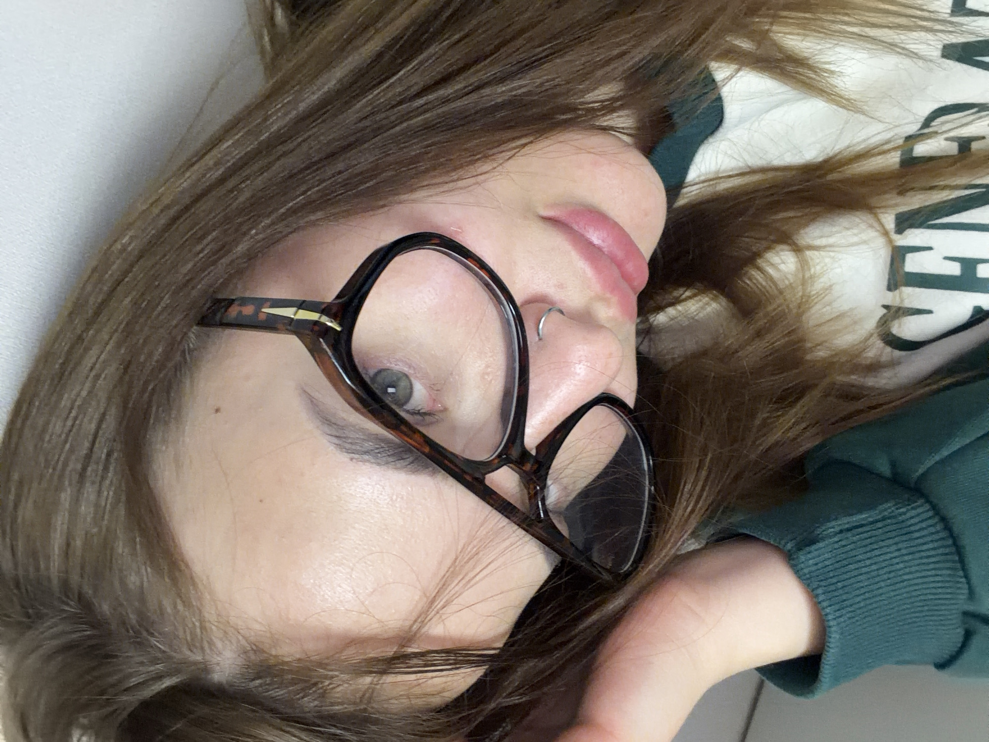 A close-up, overhead view captures a young woman lying down indoors, with the image rotated 90 degrees clockwise. She has long, light brown hair that spreads around her head and face. Her eyes are green or hazel, and she is wearing large, tortoise-shell framed glasses. A small silver ring is visible in her septum piercing. Her lips are a natural pink color and slightly parted. She is dressed in a dark green top, likely a hooded sweatshirt, with a ribbed cuff visible on her left arm. Light-colored text, possibly spelling "GENER" or "-GENERAL", is visible on the fabric of her top. Her complexion is fair. She appears to be resting on a light-colored surface, possibly a pillow or bedding, with a white background visible behind her. The pose is relaxed and candid. The provided context indicates the location is Krasnodar, Russia.