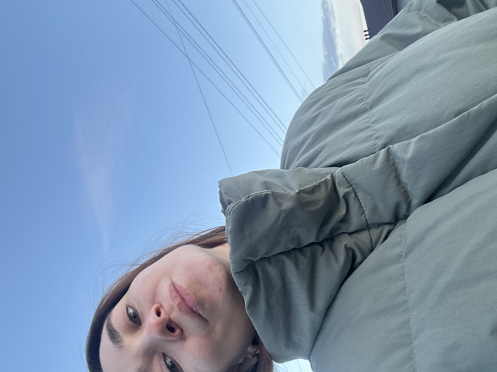 A woman in a green puffer jacket is positioned in the lower left of the frame, looking up at the camera. A clear blue sky fills the upper left portion of the image, with a set of power lines crossing diagonally. The upper right corner shows a portion of a building, possibly indicating an urban environment. No specific location information for Kharnavy, Russia is discernible.