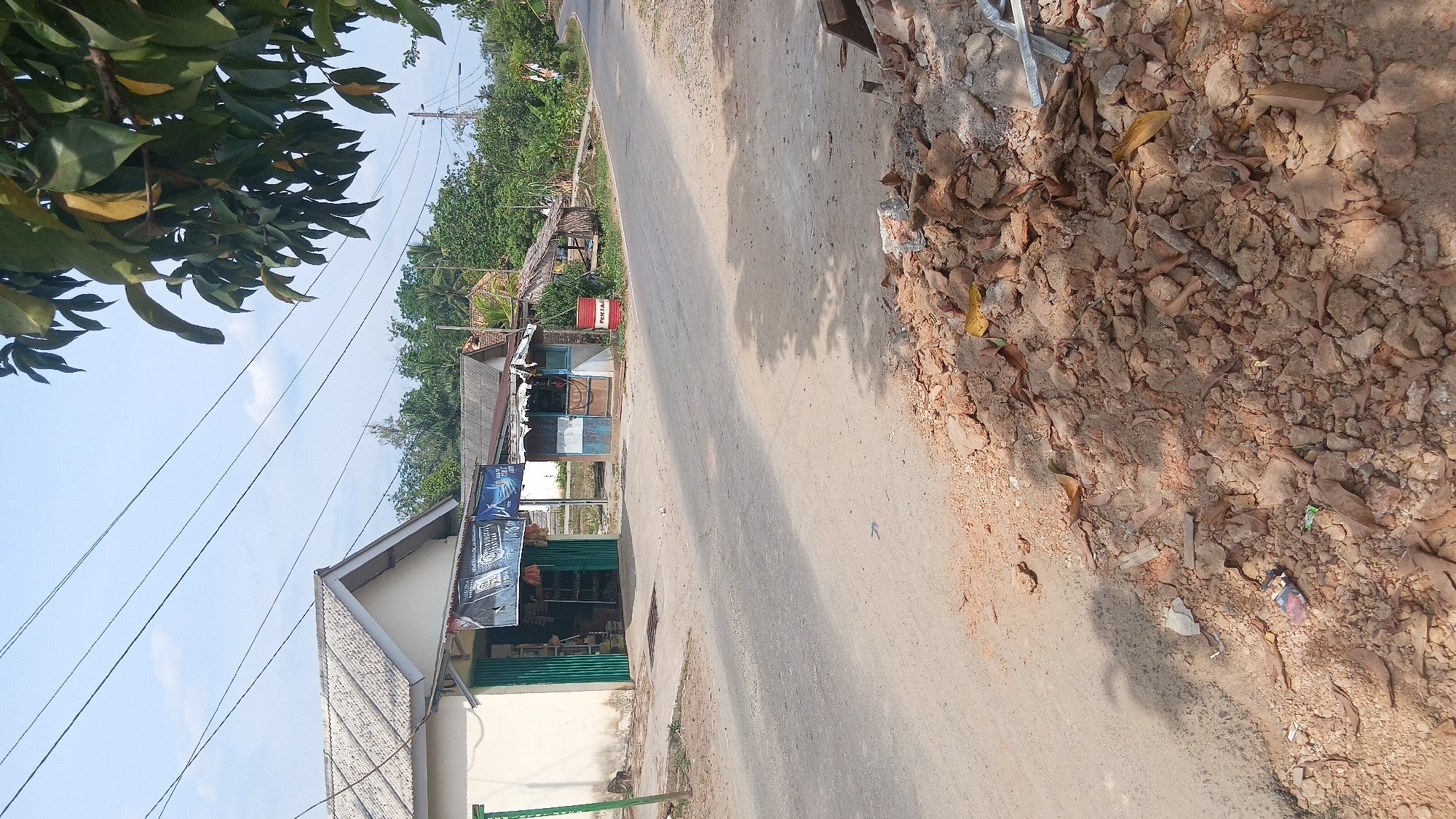 The image depicts a roadside scene in Prabumulih, Indonesia, likely a rural or semi-urban area. In the foreground, a pile of broken bricks, dirt, and dried leaves lies on the shoulder of a paved road. The road itself is somewhat worn.
Along the roadside, there are several small structures that appear to be shops or stalls. One prominent building on the left is a light-colored structure with a gray roof and green-shuttered openings, possibly a general store. Above its entrance, a banner is visible with text and graphics, suggesting it's advertising products. To the right of this building is another smaller, blue and white structure with an open front, which appears to be another shop or a small kiosk. Next to this kiosk is a red barrel with "PERTAMINA" written on it, indicating it's likely a fuel station for Pertamina, Indonesia's state-owned oil and gas company. Further to the right, there's a more rustic structure with a thatched roof.
The background consists of lush green trees and vegetation, typical of the Indonesian landscape. Overhead, power lines crisscross the sky, and a portion of a leafy tree is visible in the upper right corner of the frame. The sky is clear with some light clouds. The overall impression is one of a modest, functional roadside environment serving the local community.