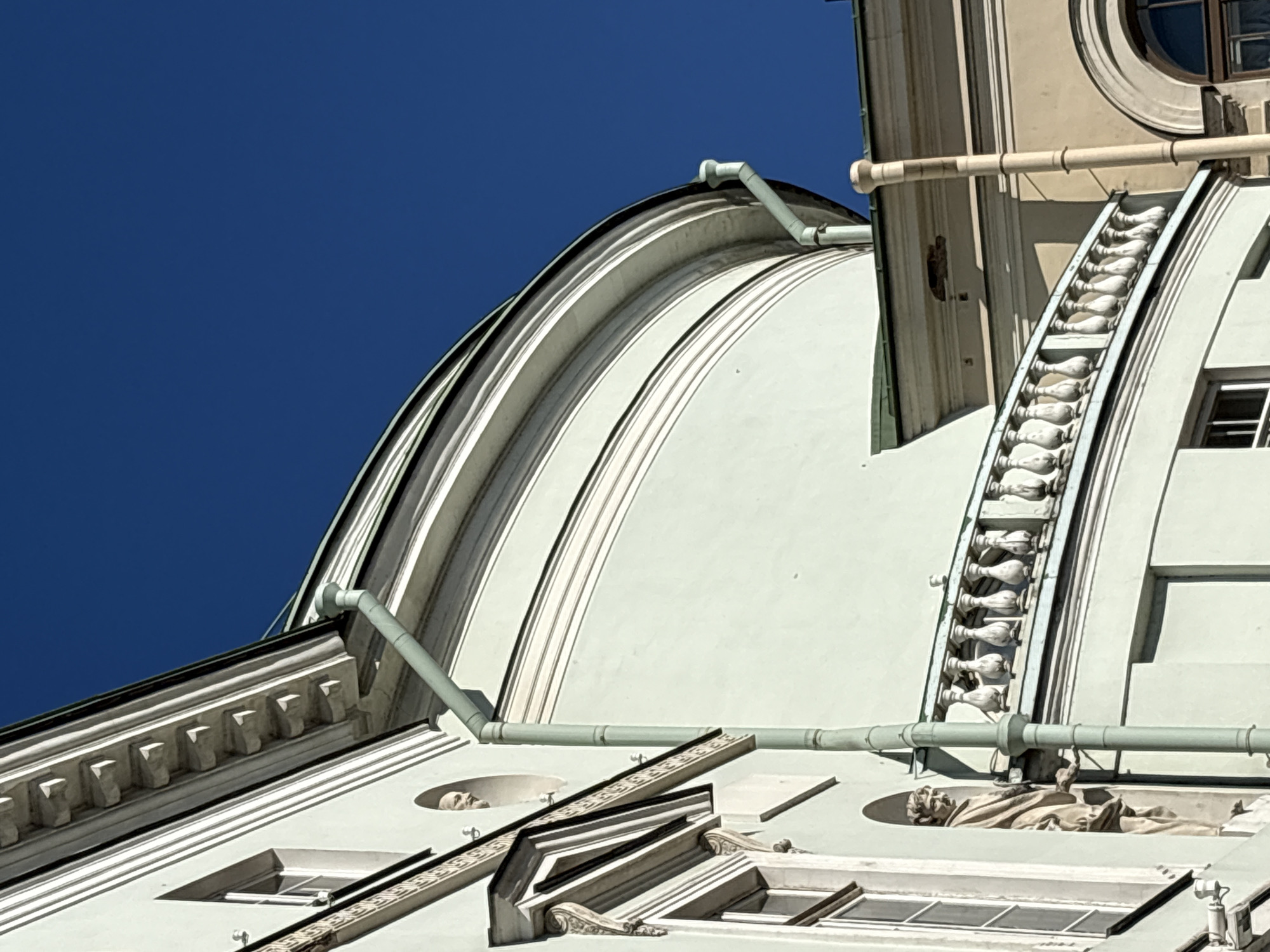An upward-angled view captures the architectural details of a building against a clear, deep blue sky. The primary facade is a pale green-grey, featuring prominent white horizontal banding and cornices. On the left, a rectangular window is visible, below which a full-figure classical statue is recessed within a niche. Further up, a circular opening contains a classical male bust. A segmented, pale green downspout descends vertically along this section of the facade. The building curves to the right, culminating in a large, smooth dome structure with a visible dark green roofline and multiple white horizontal stripes. Below the dome, a white balustrade, comprising several classical balusters, follows the building's curvature. To the far right, a section of an adjacent or connected building, rendered in beige, is partially visible, displaying an arched window and a separate, cream-colored downspout. The scene is under bright daylight, creating distinct shadows. Minor surface wear and discoloration are present on the building's exterior, including a small area of material loss on the beige section's roofline. No people are depicted. The location is in the city of Unknown, Unknown.