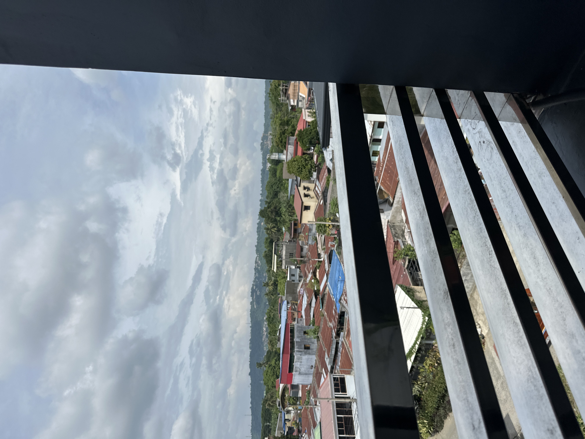 View from a high vantage point, possibly a building in Davao City, Philippines. Predominantly gray skies filled with clouds are overhead. Below, a residential area with houses with various colored roofs, predominantly red and blue, interspersed with green trees. A gray metal railing is in the foreground, partially obscuring the view.