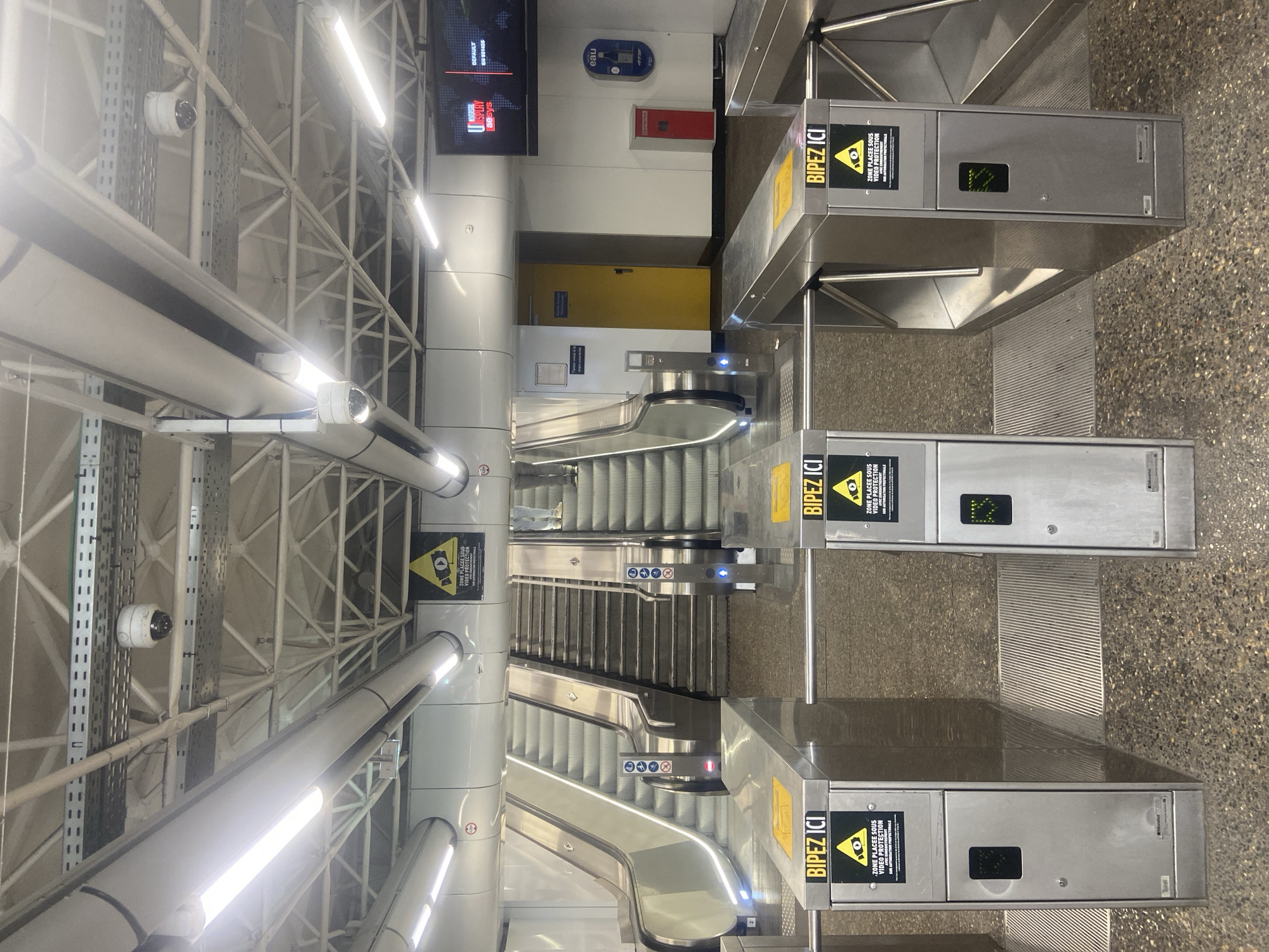 The image depicts a subway station turnstile area. In the foreground, three stainless steel turnstiles are visible, each with a yellow sign at the top displaying the text "BIPEZ ICI" which translates to "Scan Here." Below this, a triangular yellow warning sign with an eye symbol indicates video surveillance. The turnstiles have small black rectangular screens or indicators and a keyed lock on the lower front panel. The floor is a speckled granite material.
In the middle ground, a set of escalators and stairs lead upwards. The escalators are stainless steel, and the stairs are concrete. Two security cameras are visible mounted on the ceiling structure above the escalators. Further back, a bright yellow door is set into a white wall. To the right of the door, a red fire alarm or extinguisher box is mounted on the wall, and an "eau" (water) dispenser is also present. A large monitor displaying "DISPLAYS ae.sys" is mounted high on the wall.
In the background, the ceiling is an intricate network of white metal beams and supports, with extensive conduit and lighting fixtures. Several white, dome-shaped security cameras are mounted on the ceiling. The overall environment suggests a modern, functional public transportation hub.