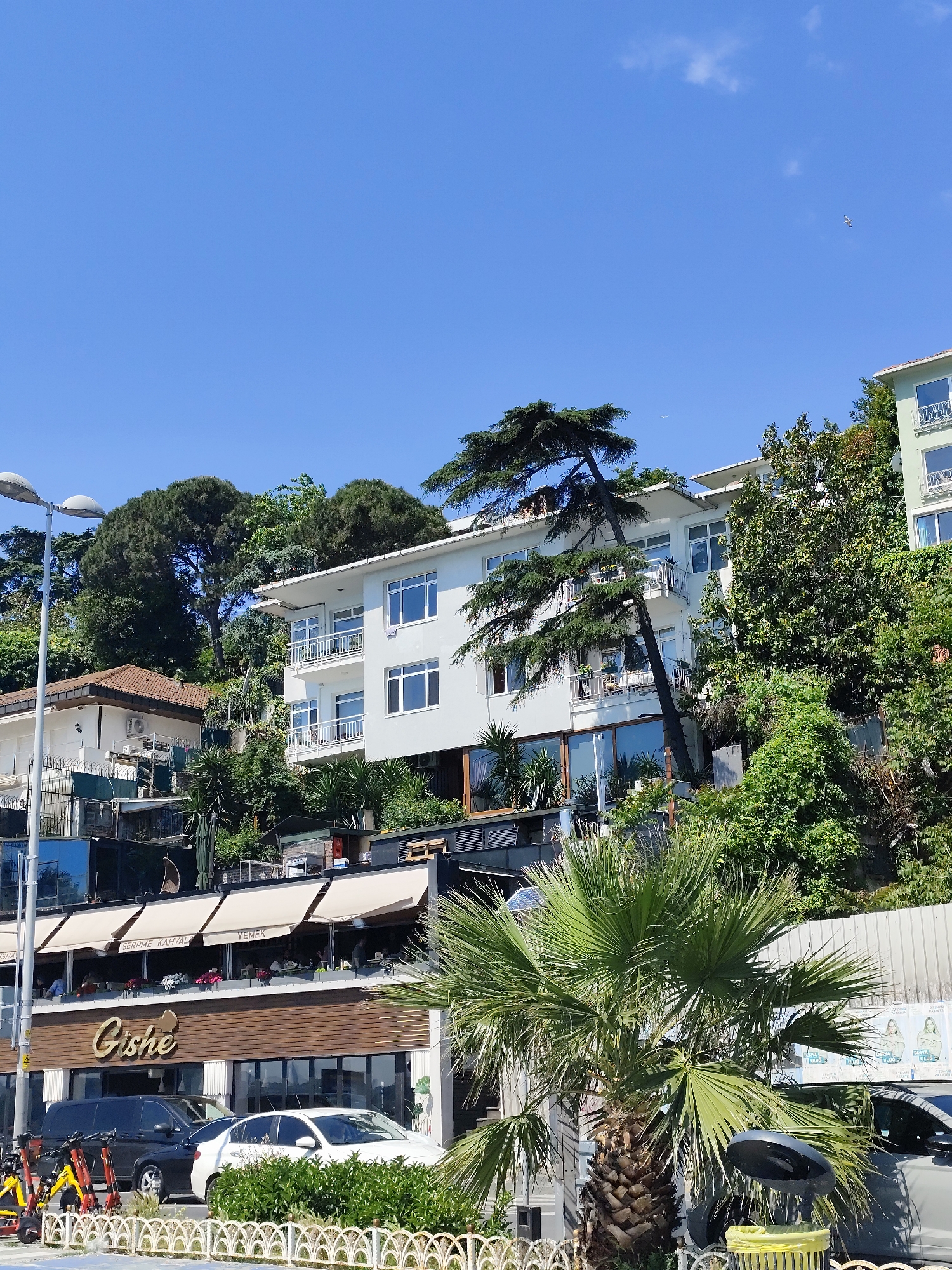 Captured in Sarıyer, Turkey, the image presents an outdoor scene under a clear blue sky. The architecture features multi-level buildings with a blend of residential and commercial spaces. The "Gishe" restaurant stands out with its wooden facade and awning-covered terrace where people are seated. A variety of vehicles, including cars and scooters, are parked in front of the establishment. Lush greenery, including a palm tree, surrounds the buildings, which are constructed on a hillside. A single bird can be spotted in the sky.