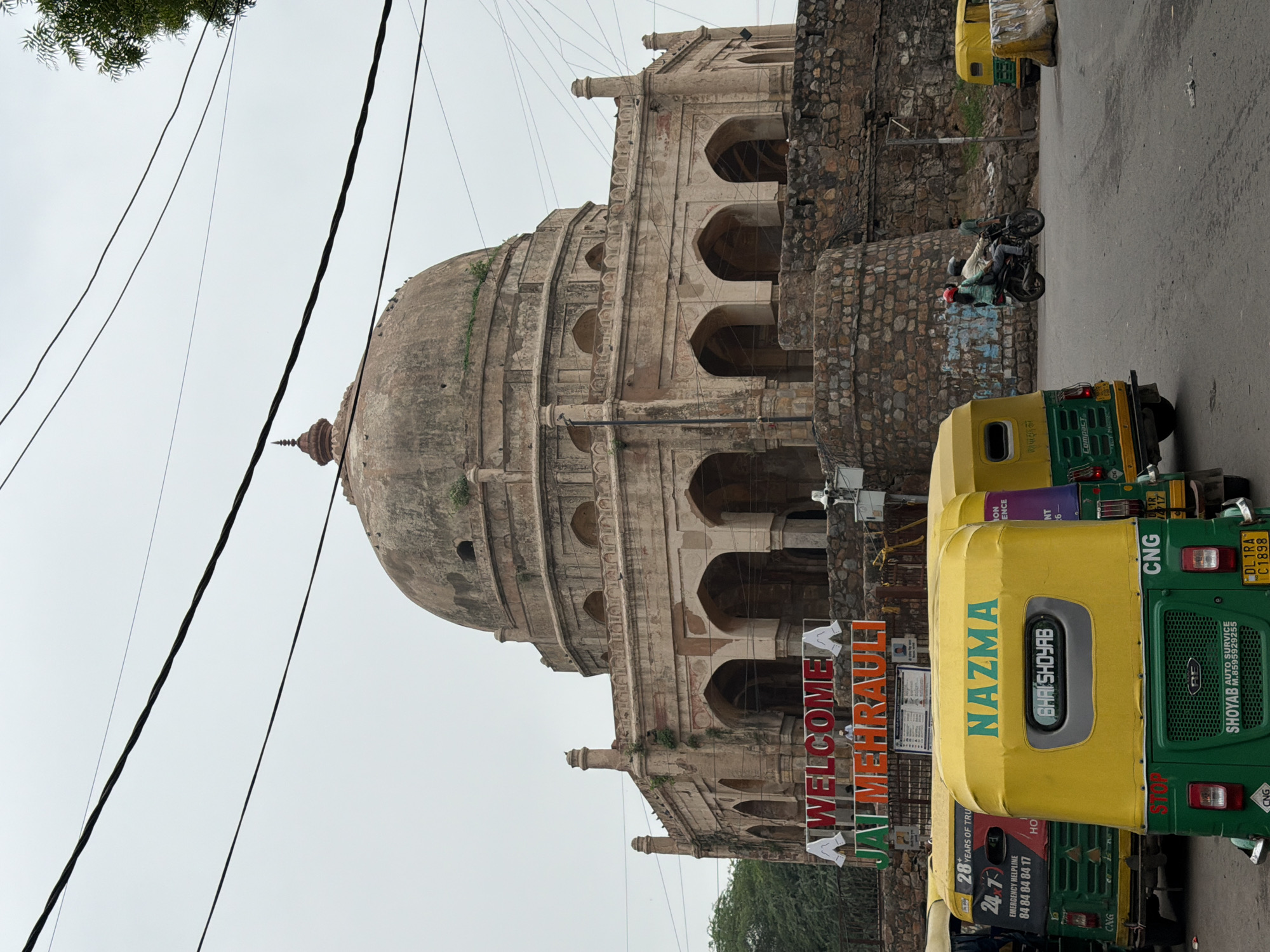 The image depicts a street scene in Mehrauli, a historical area in Delhi, India. In the background, a large, domed, ancient stone structure, likely a monument or tomb, stands under an overcast sky. The structure features arched entrances on its lower level and is partially covered in greenery. In the foreground, several yellow and green auto-rickshaws are parked on a paved road. One auto-rickshaw has a sign that reads "WELCOME JAI MEHRAULI" in red and green lettering. Another auto-rickshaw displays "NAZMA" in green text on its yellow roof. Two people are visible on a motorcycle, with the rider wearing a helmet and a passenger wearing a red helmet. A third person stands nearby. Electrical wires are strung across the upper portion of the image. The overall scene suggests a typical day in a busy urban environment with historical significance.