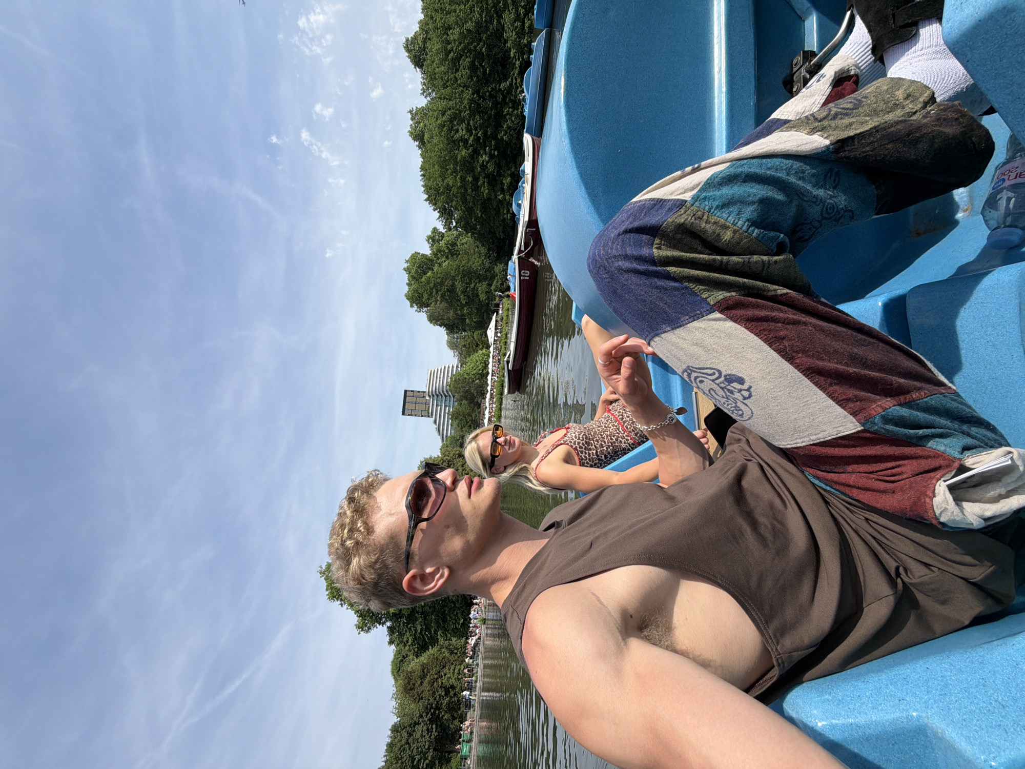 A medium eye-level shot shows a person in a brown tank top and patchwork pants sitting in a blue paddleboat in a lake, with another person in a leopard-print top visible in the background. The sky is blue with some scattered clouds. The location is in a park in London, United Kingdom, as there is a modern building resembling a skyscraper in the background. Trees and other greenery surround the lake. There are people sitting along the edge of the lake. The focus is on the person in the foreground, with the other person and the background slightly blurred. A water bottle sits to the right of the patchwork pants.
