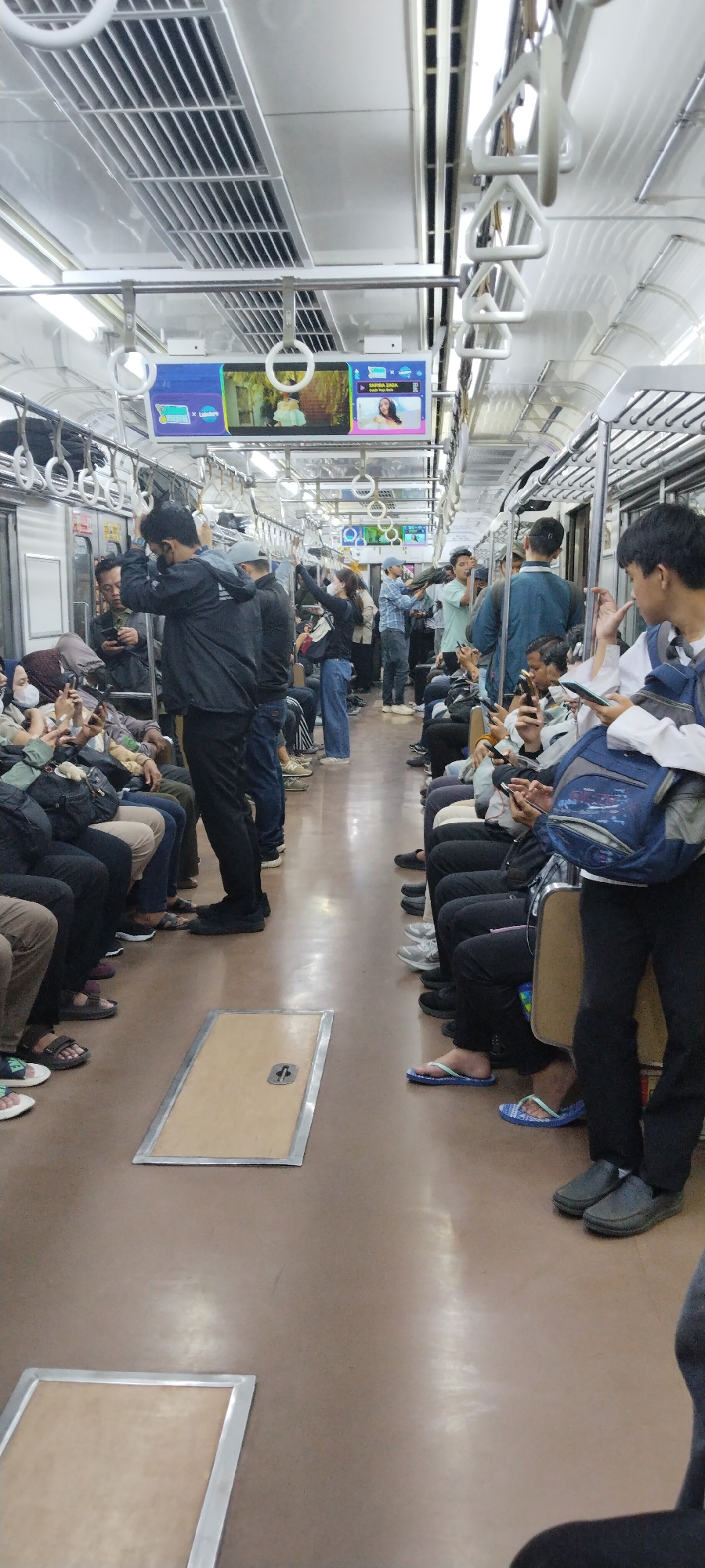 The image depicts the interior of a crowded train carriage, likely a commuter train, with many passengers standing and sitting. The overall environment is utilitarian, with rows of overhead handles for standing passengers and what appear to be bench seats along the sides. The floor is a neutral, brown-toned material. There are digital screens mounted above the windows displaying advertisements or information, with one screen showing a woman in a bright pink top and another showing a scene with trees and a person.
Several individuals are the primary subjects. In the foreground on the right, a young man with dark hair is looking down at his phone, wearing a white shirt and a blue backpack. To his left, a person in dark pants is wearing grey slip-on shoes. Further down the carriage, a woman in a black top and blue jeans is standing and looking upwards, her hand raised. There are many other passengers, most of whom are holding or looking at their mobile phones, indicating widespread use of personal devices. Some individuals are wearing face masks. The lighting appears to be from overhead fluorescent fixtures.
The scene suggests a typical urban commuting experience in a populated city. While the image itself does not contain explicit markers of Central Jakarta, Indonesia, the style of the train, the demographic of the people, and the ubiquitous use of smartphones are consistent with transportation environments in that region. The presence of advertisements on screens further supports an urban commercial setting. The mood is one of passive activity, with most individuals engaged with their personal devices rather than interacting with each other.