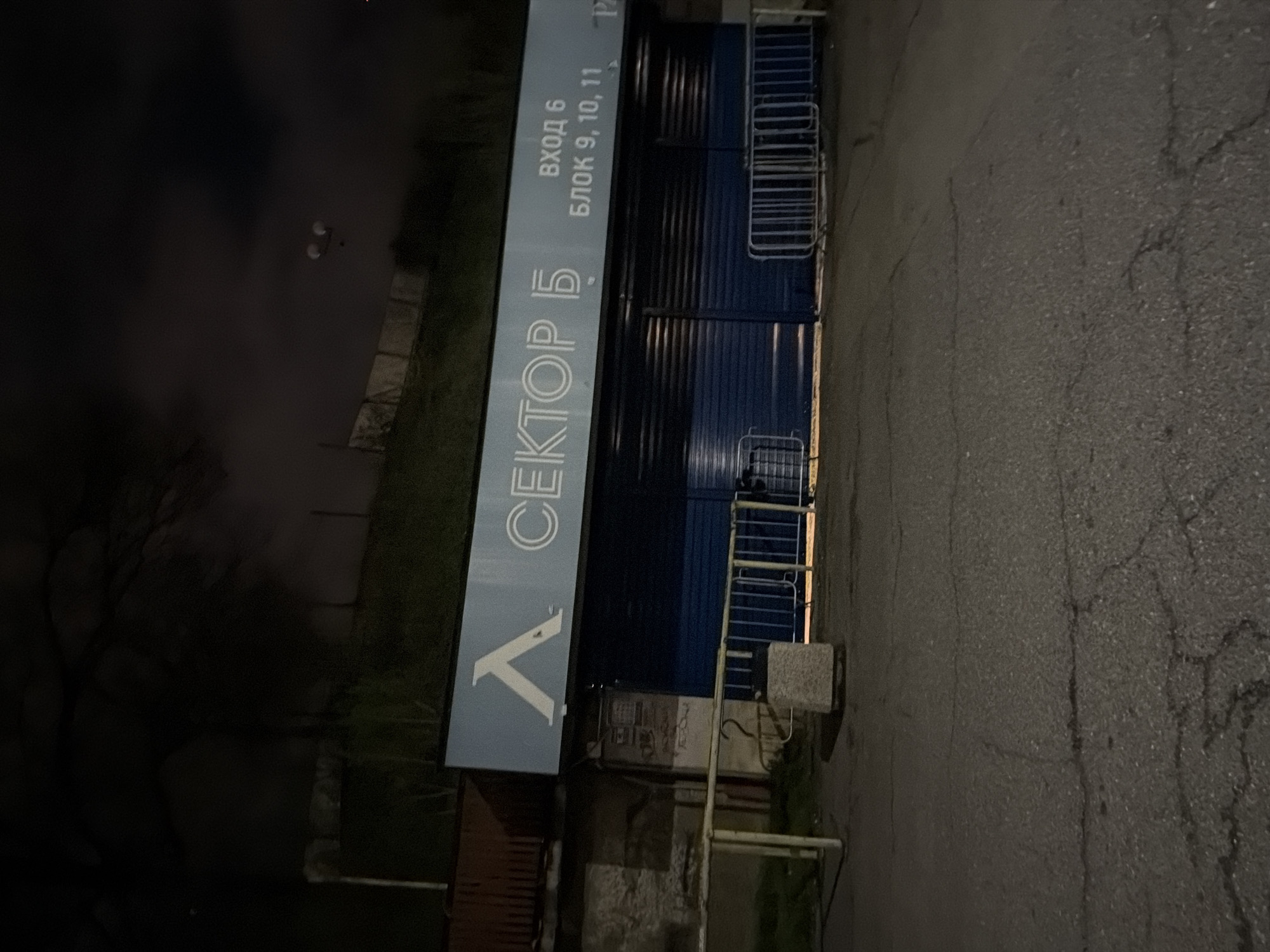 The image depicts the exterior of a building at night. A vertical sign reads "СЕКТОР Б" and "ВХОД 6 БЛОК 9, 10, 11". The building itself is a dark blue structure with corrugated metal siding. There are metal staircases leading to higher levels. The ground is asphalt and appears cracked. The surrounding environment includes some trees and vegetation in shadow. The location is likely in Sofia, Bulgaria based on the Cyrillic text on the sign.