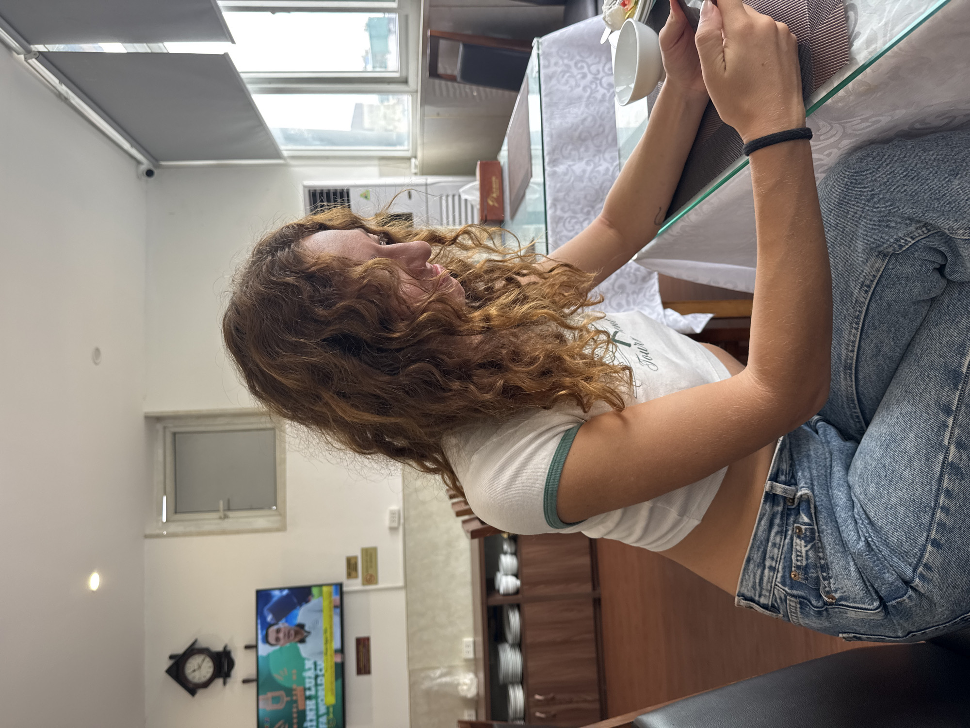 In a room that appears to be a dining area, possibly in Hà Nội, Vietnam, a young woman with curly auburn hair is seated at a glass-topped table, looking down at a mobile device in her hands. She is wearing a white crop top with a green trim and light blue denim jeans. Her left arm, adorned with a black braided bracelet, rests on the table. A white bowl and a plate with food are visible on the table in front of her. The room features a dark wooden cabinet displaying stacks of white plates and bowls, and a television mounted on the wall playing a program with Vietnamese text and an image of a man. Other elements in the scene include a black cuckoo clock on the wall, grey roller blinds covering a window, and a security camera visible on the ceiling. The flooring is dark wood.