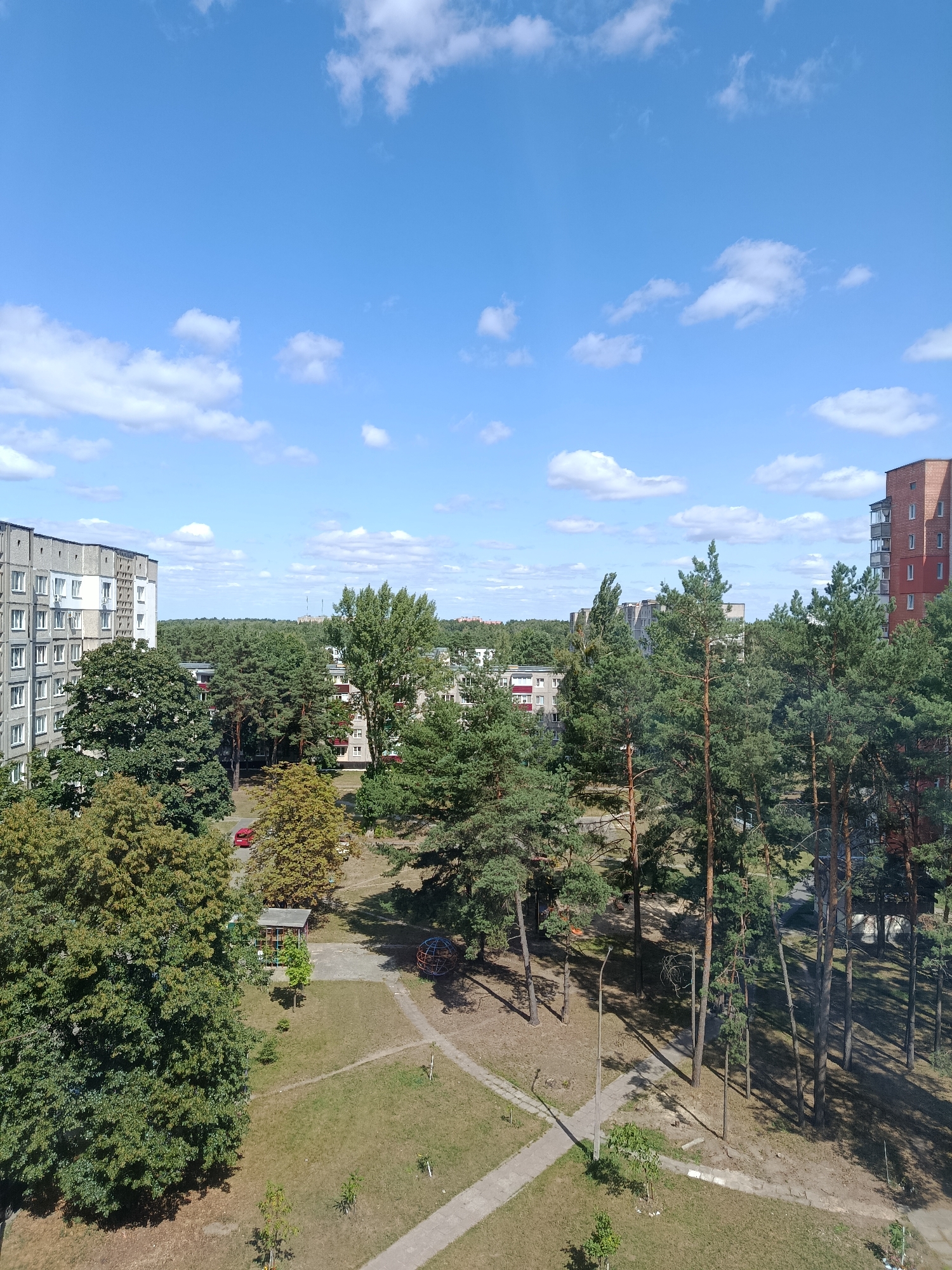 This is a daytime, medium-angle, full shot of a residential area in Svietlahorsk, Belarus. The sky is bright blue with scattered white cumulus clouds. In the foreground and midground, there are numerous tall pine trees with dense green foliage. To the left, a multi-story concrete apartment building with numerous windows dominates the scene. Further back, more apartment buildings of similar design are visible, interspersed with more trees. A red car is parked near one of the buildings. In the central part of the image, there is a playground with a climbing structure and a small gazebo-like shelter. A concrete pathway winds through the grassy courtyard, bordered by dry grass and scattered small trees. The scene depicts a typical urban environment with significant green space.